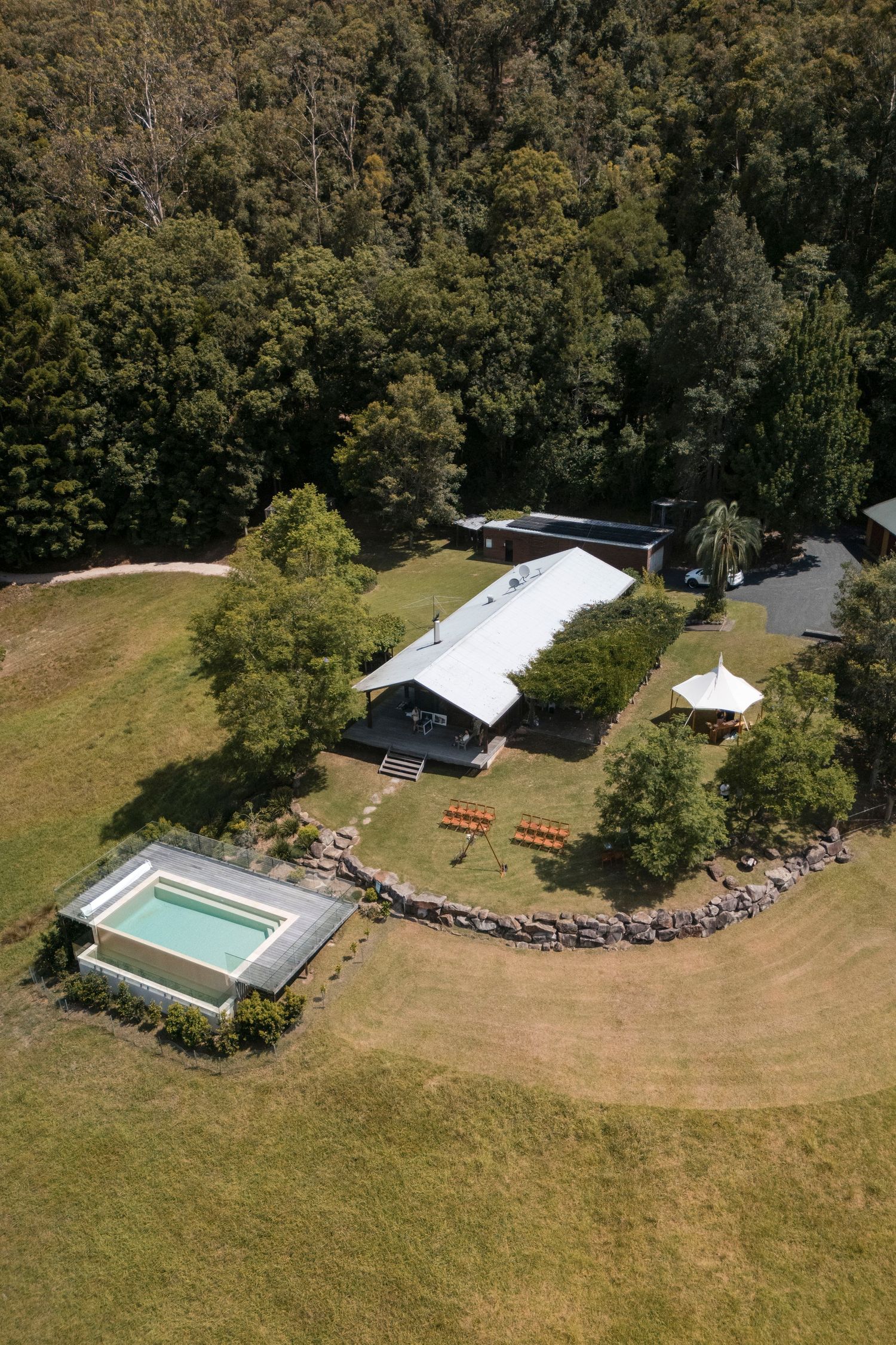 Aerial view of a property with swimming pool, white tent, and buildings nestled in a forested hillside.