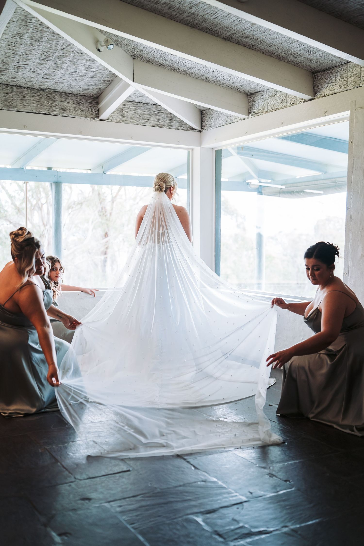 Bridesmaids help arrange flowing white wedding veil on bride in sunlit room with blue-tinted windows.