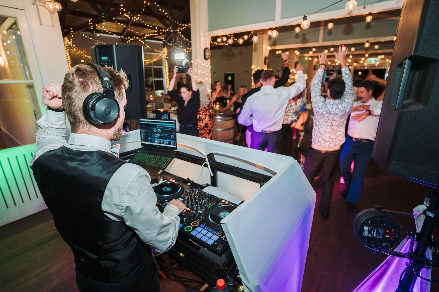 DJ in formal attire plays music at a wedding reception while guests dance on the illuminated dance floor.