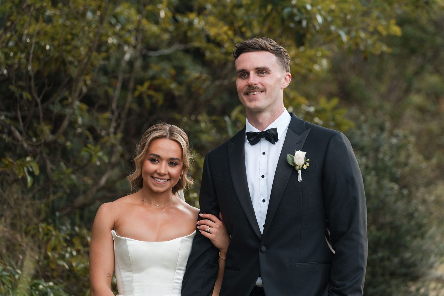 Bride and groom posing outdoors in wedding attire, she in white strapless gown, he in black tuxedo with boutonniere.