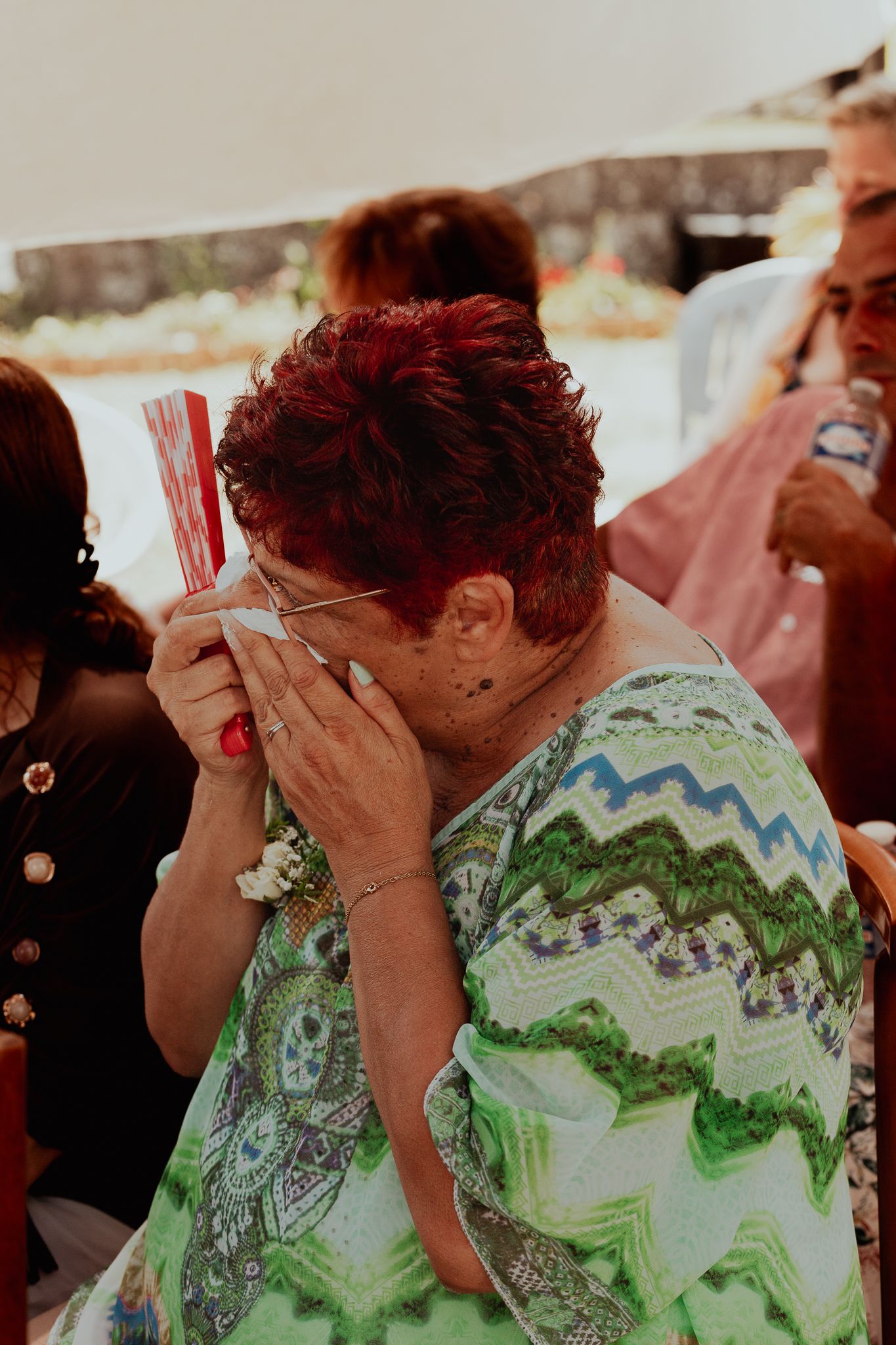 Emotional woman with red hair covering her face at an outdoor event, wearing a colorful green patterned dress.