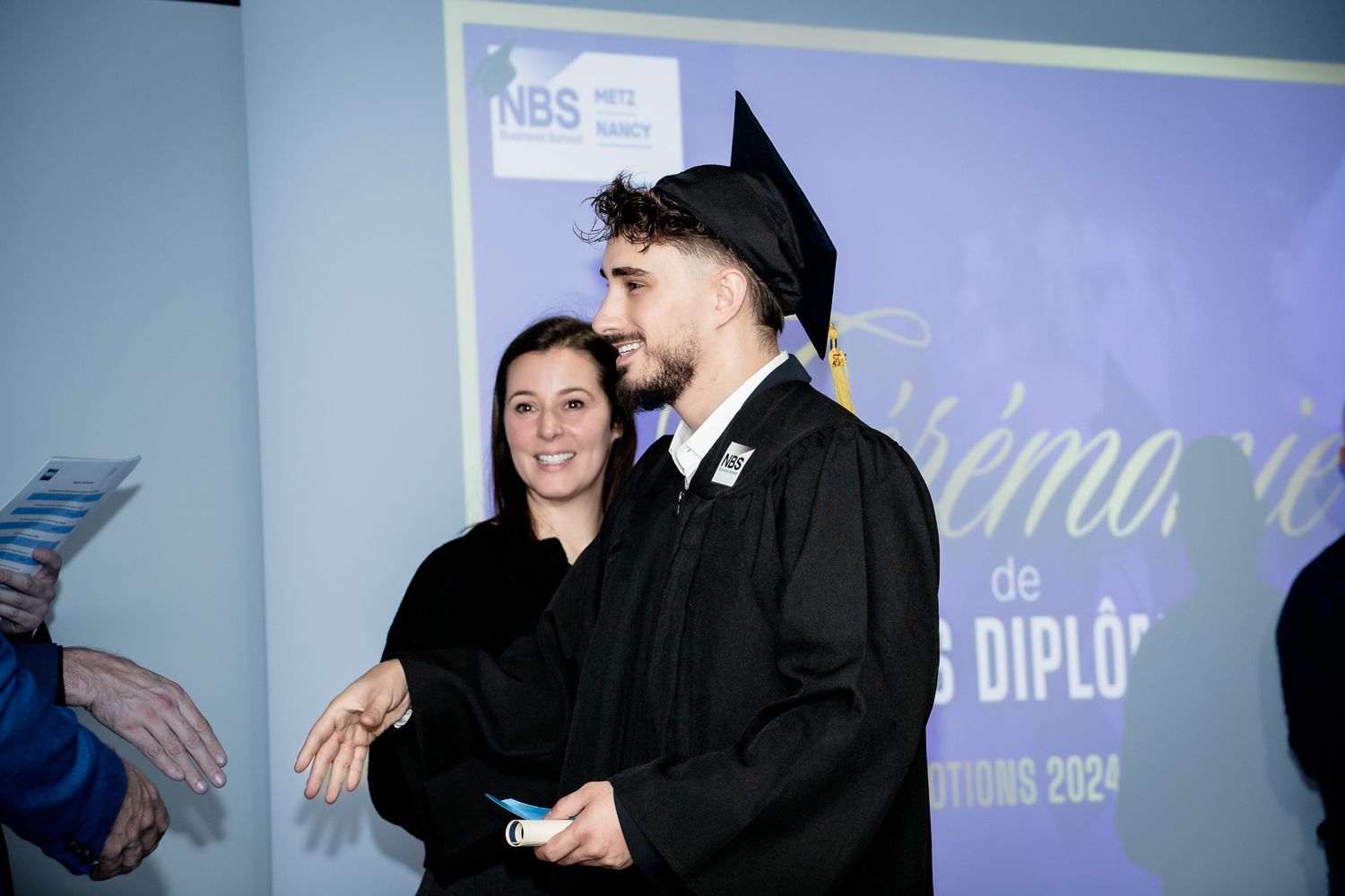 Graduate in cap and gown posing with family member at NBS ceremony.