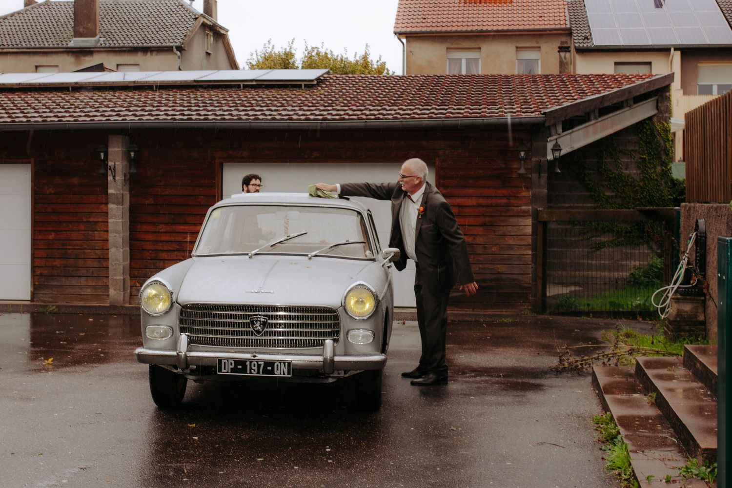 Elderly man standing beside a vintage white Peugeot 404 car parked on a wet driveway outside a wooden garage.