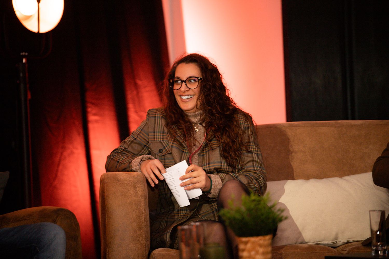 Smiling woman with curly hair and glasses seated on sofa during a panel discussion on stage.