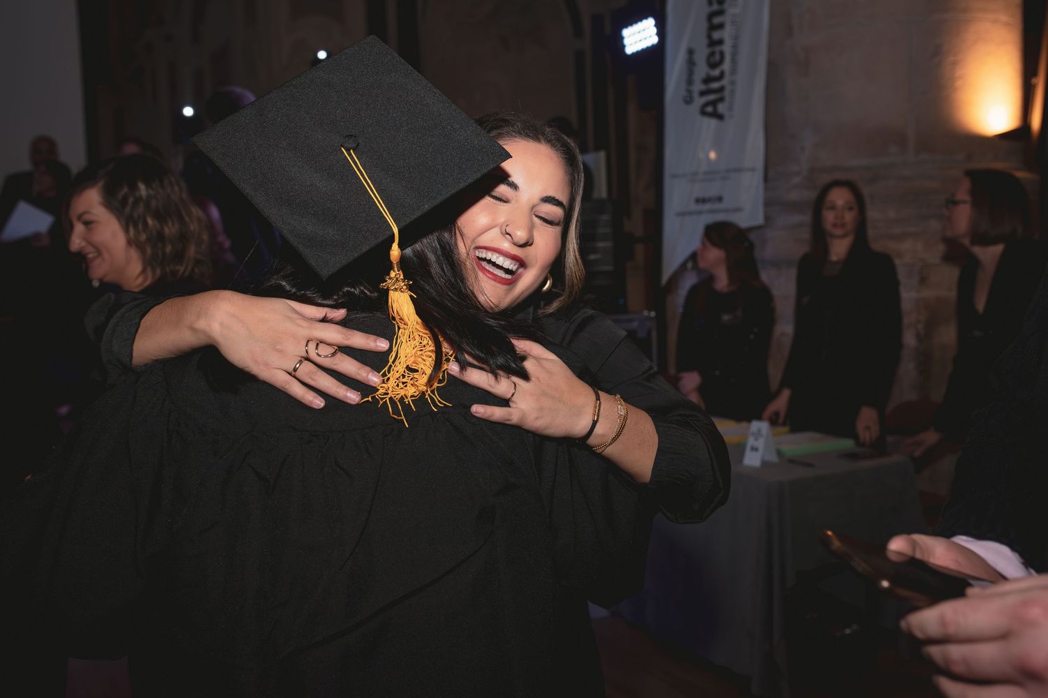 Two graduates in black caps and gowns sharing a warm hug at graduation.