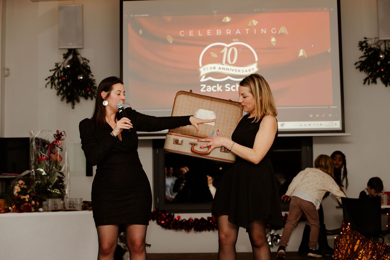 Two women in black dresses present a vintage suitcase on stage at a 10th anniversary celebration.