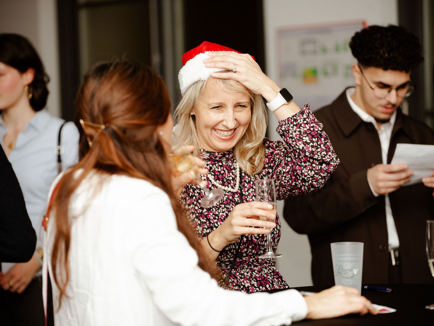 A smiling blonde woman wearing a Santa hat enjoys a drink at a festive Christmas party.