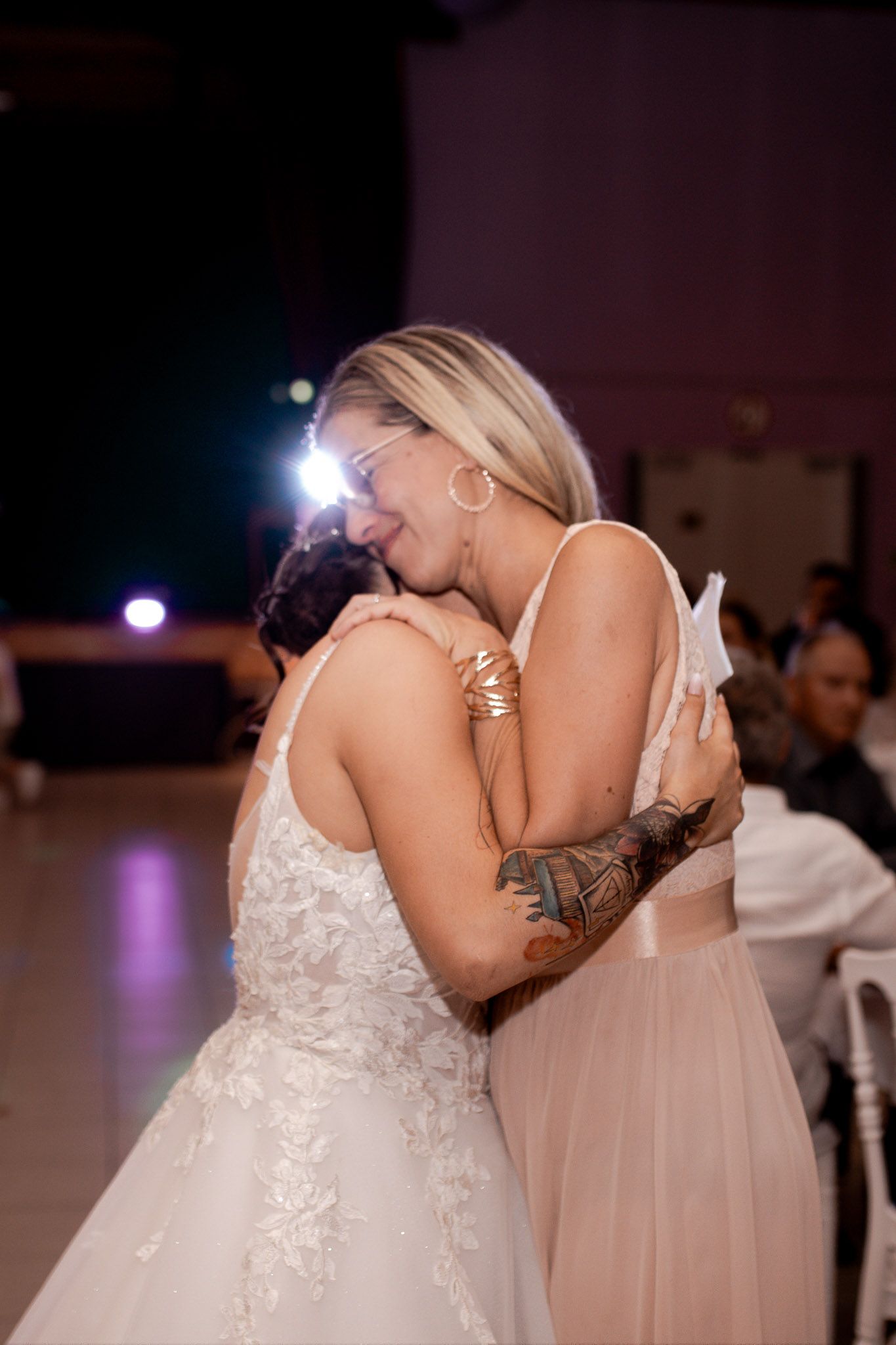 Bride and a tattooed guest share an emotional hug on the dance floor at a wedding reception.