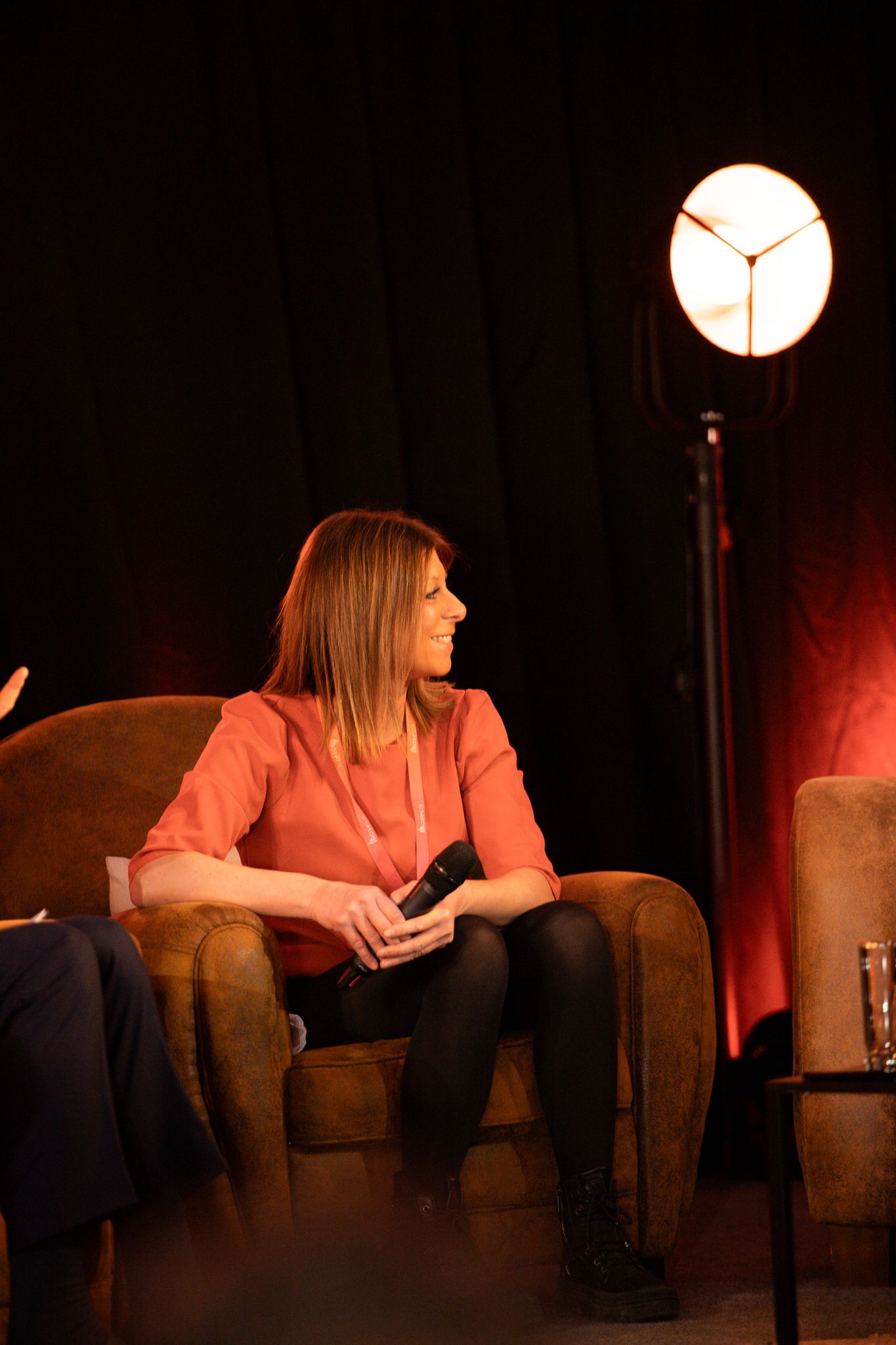Woman in coral blouse seated in armchair on stage under warm spotlight at a panel event.