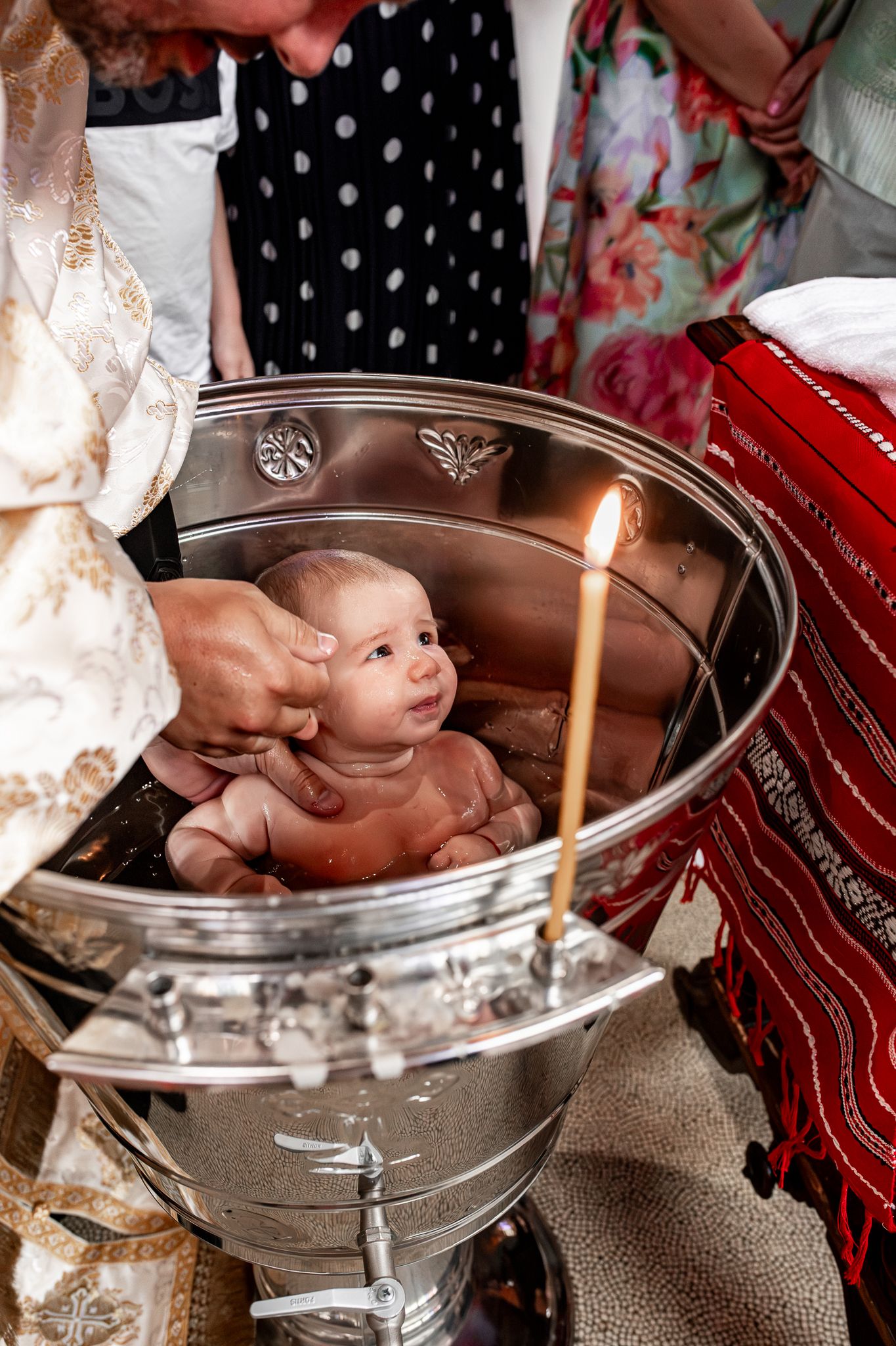 Newborn baby being baptized in a silver font with a lit candle during ceremony.