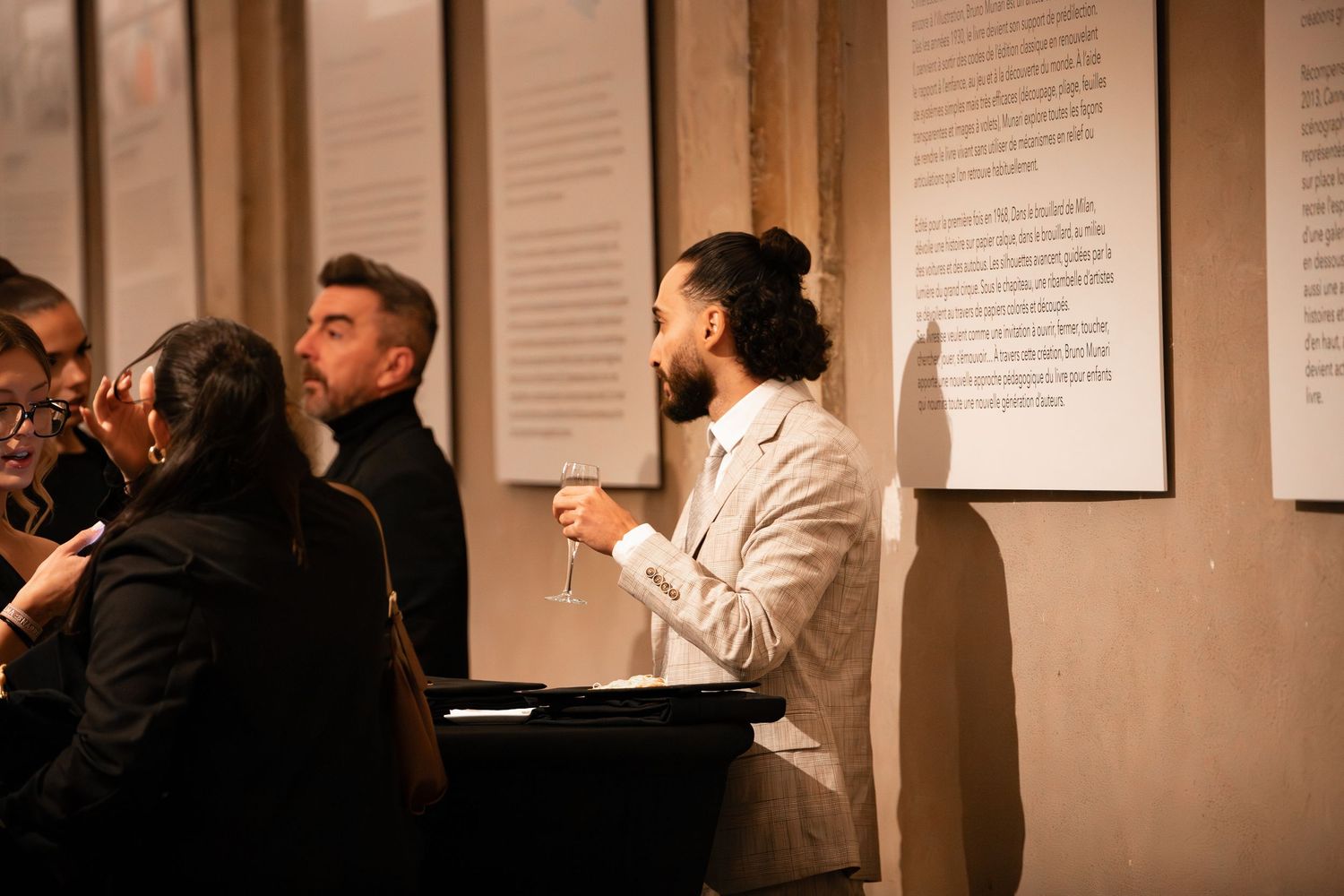 Man in beige jacket holding wine glass viewing exhibition wall text.