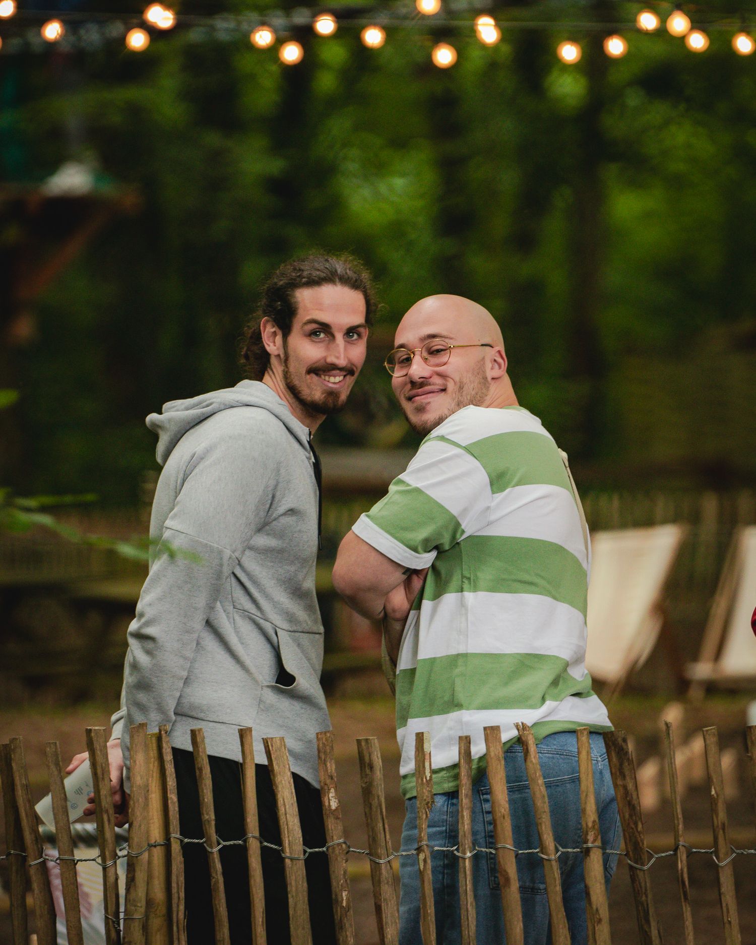 Two smiling men pose together near a wooden fence in a lush green outdoor setting.