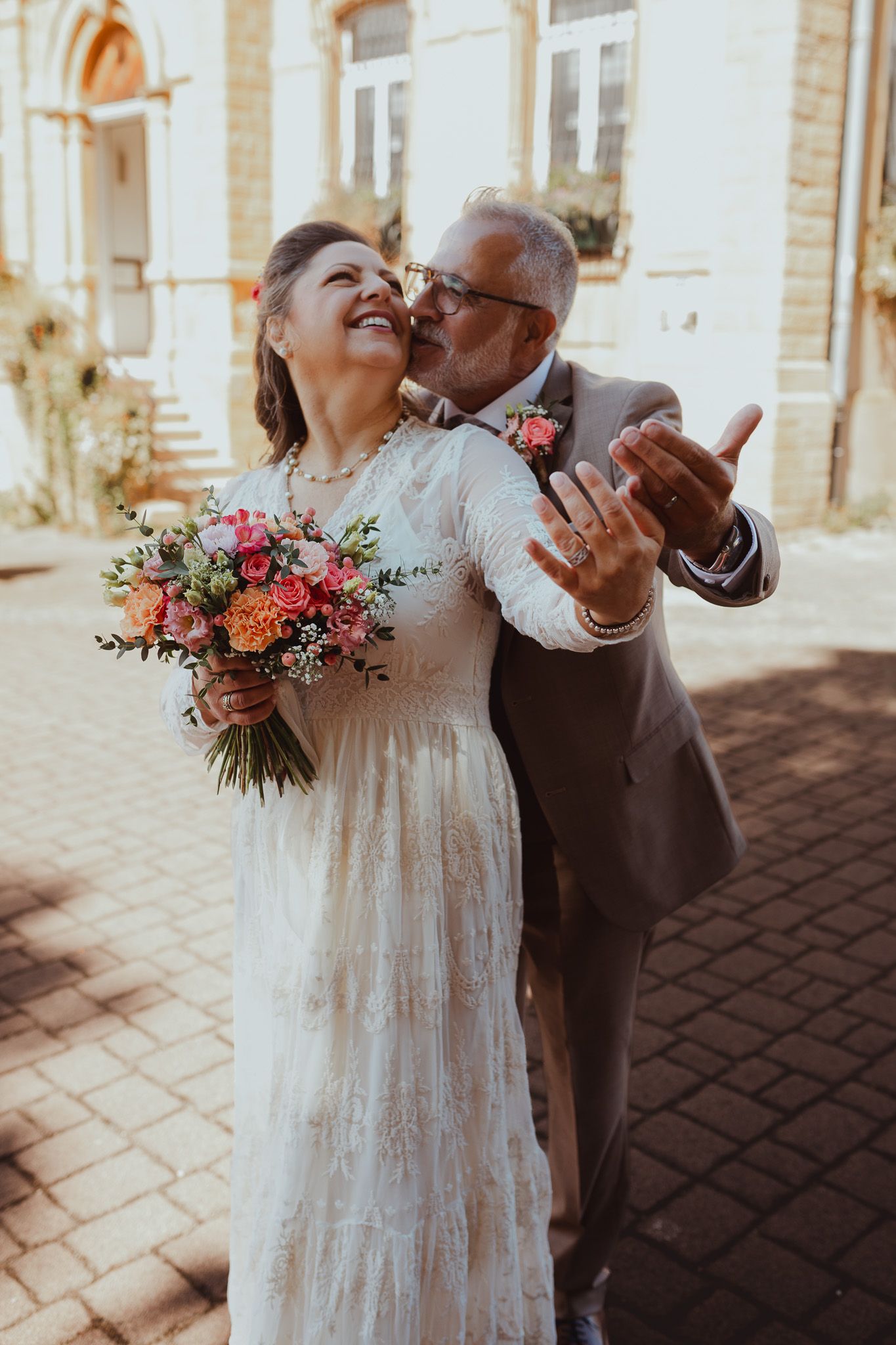 Joyeuse mariée en robe en dentelle tenant un bouquet coloré alors que le marié lui embrasse la joue à l'extérieur.