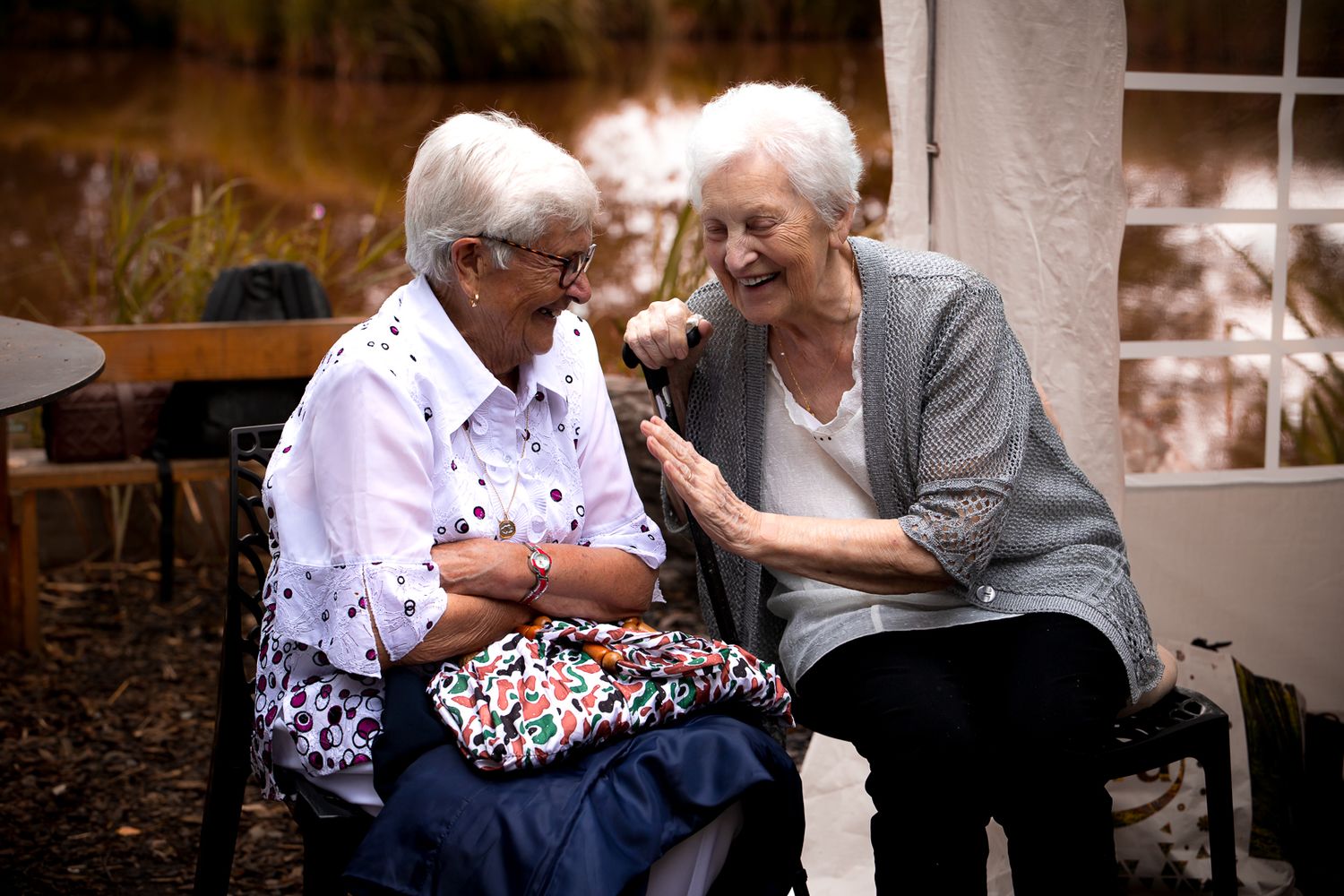 Two elderly women sharing a joyful moment outdoors, laughing and holding hands in a garden setting.