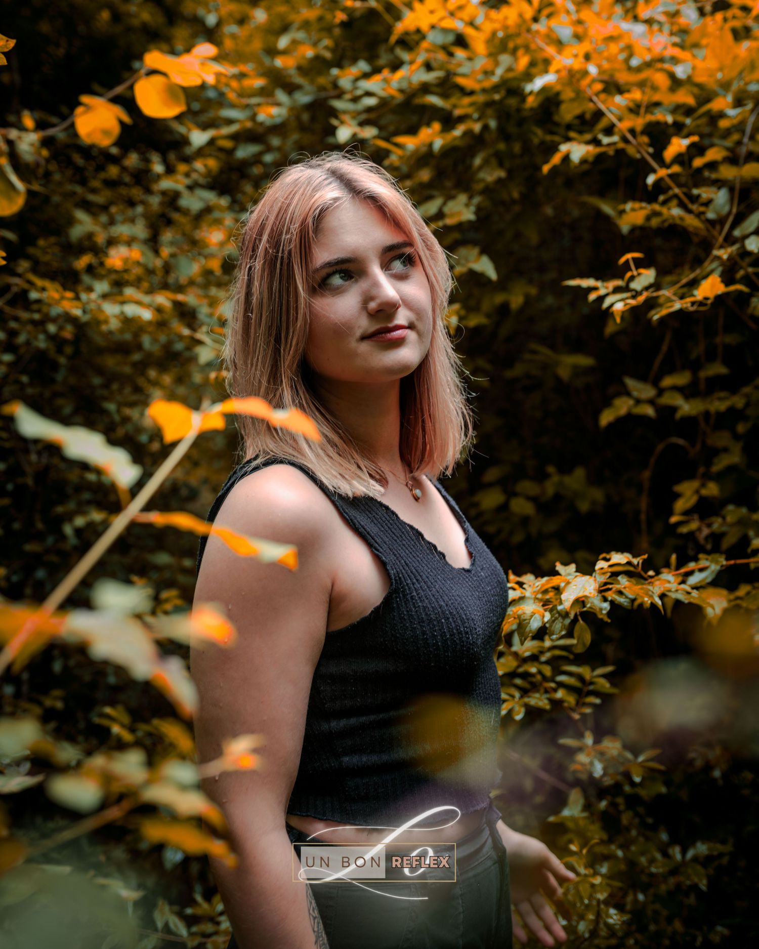 Young woman with pink-tinted blonde hair poses among vibrant autumn foliage in a black sleeveless top.