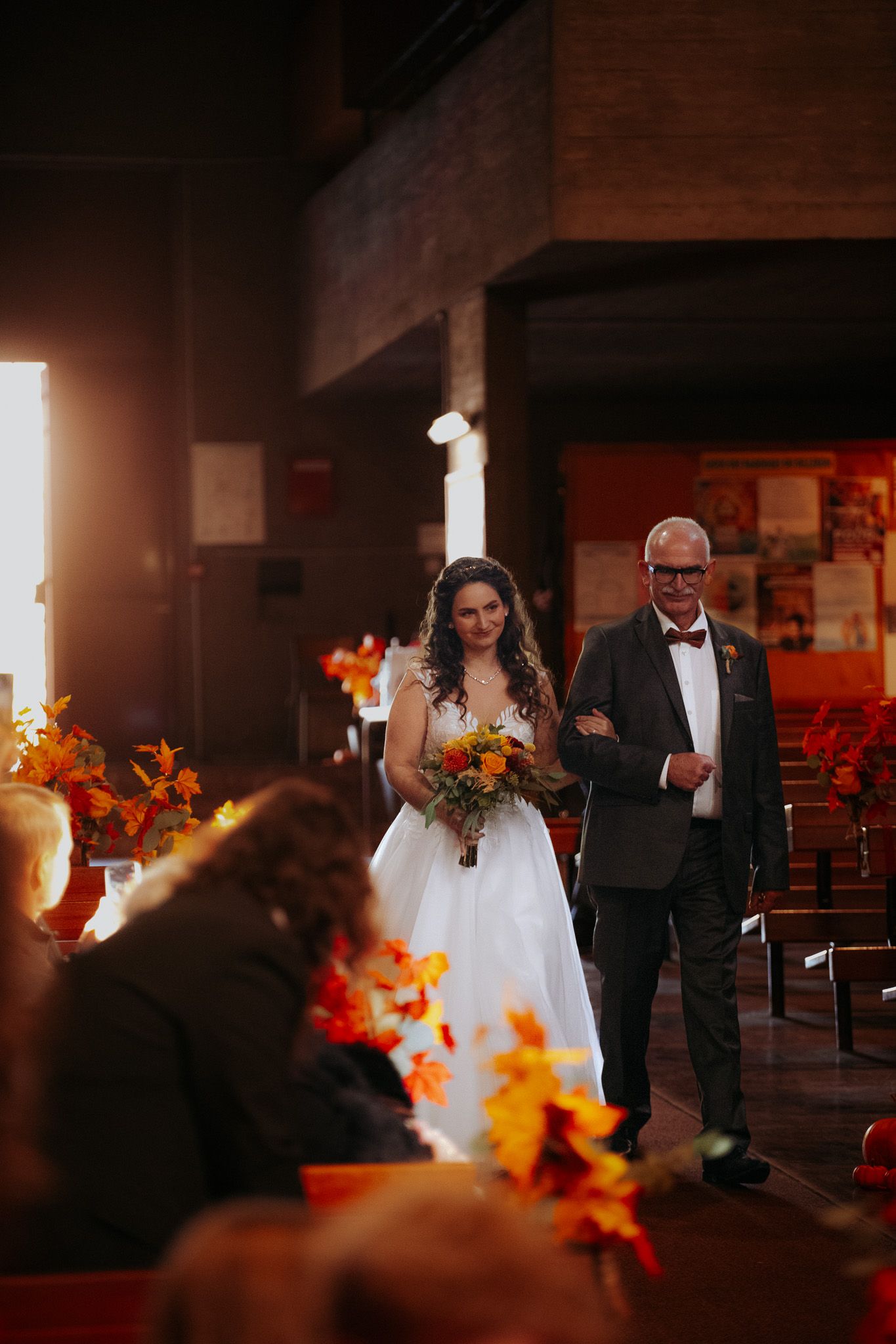 Bride walks down the aisle with her father amid warm golden light and autumn floral decorations.