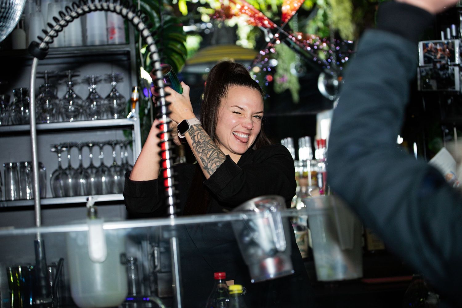Smiling tattooed bartender laughing behind the bar at a busy nightclub event.