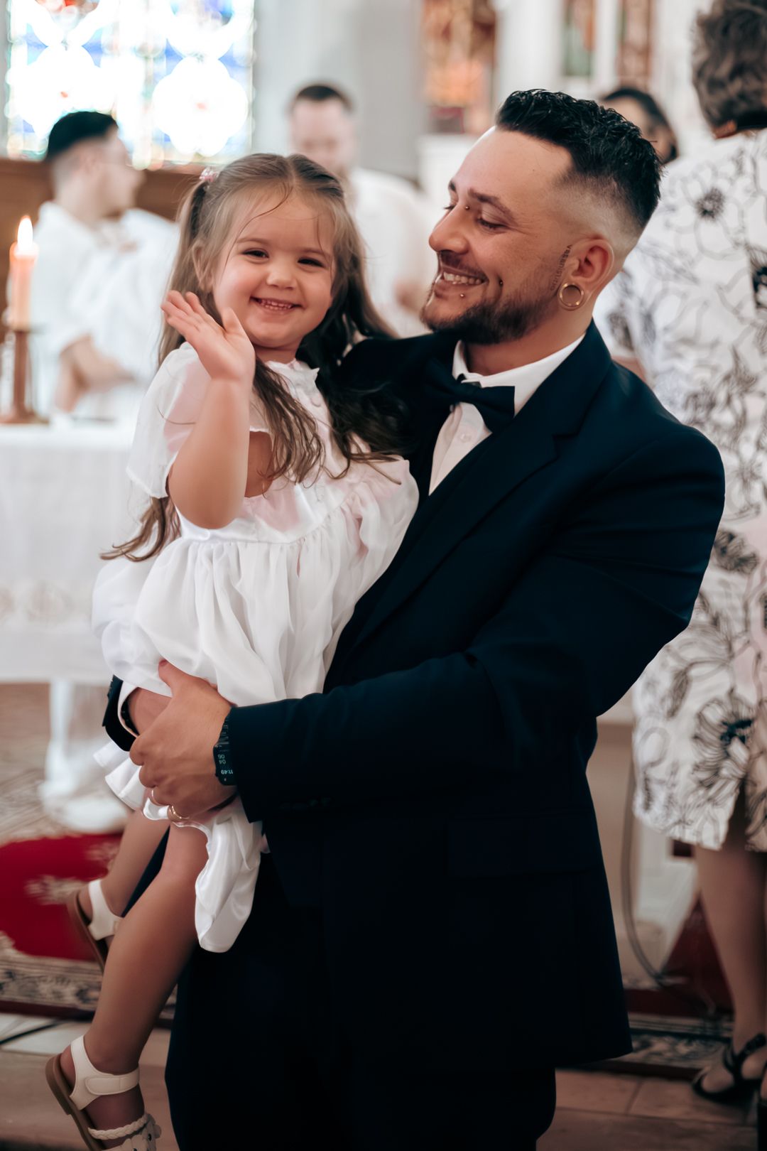 Smiling father in tuxedo holding happy little girl in white dress at church ceremony.