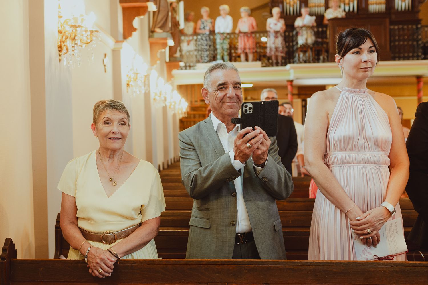 Three wedding guests standing in elegant church, man applauding between two women.