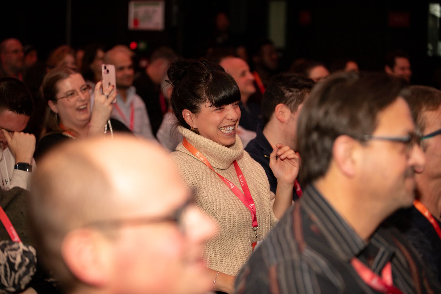 Enthusiastic woman laughing and taking photos in a crowded conference audience wearing an orange lanyard.