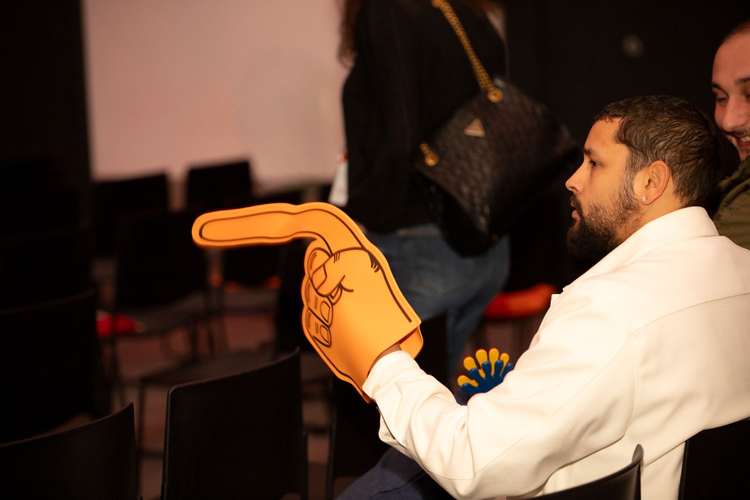Homme en veste blanche tenant un grand doigt en mousse lors d'un événement intérieur dans un auditorium sombre.