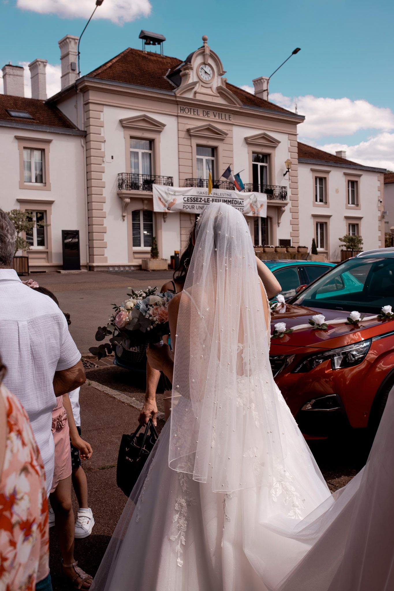 Bride in white wedding gown and veil walks toward a French town hall on a sunny day.