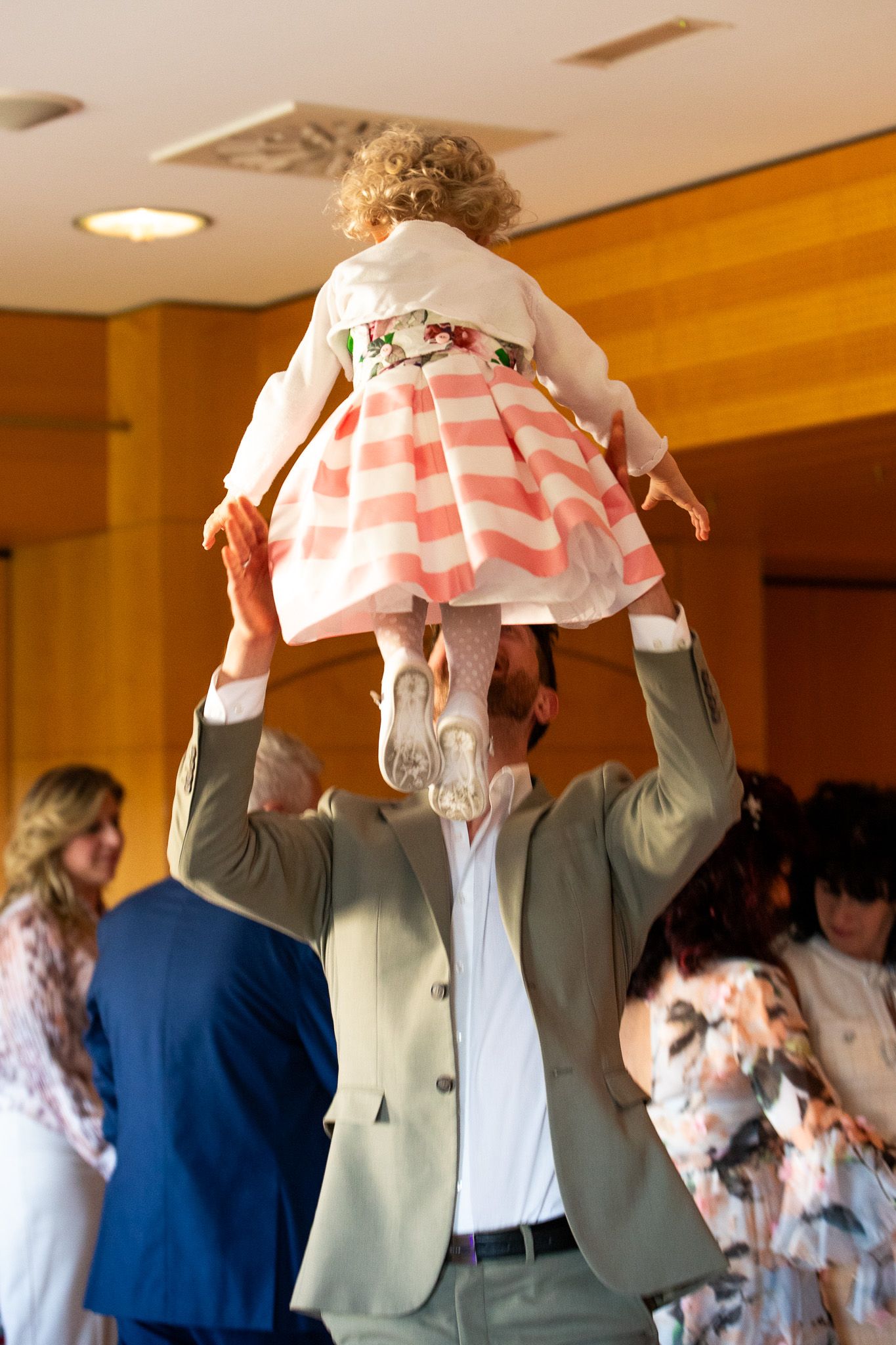 Man in suit joyfully lifting a curly-haired toddler girl in pink striped dress above his head.