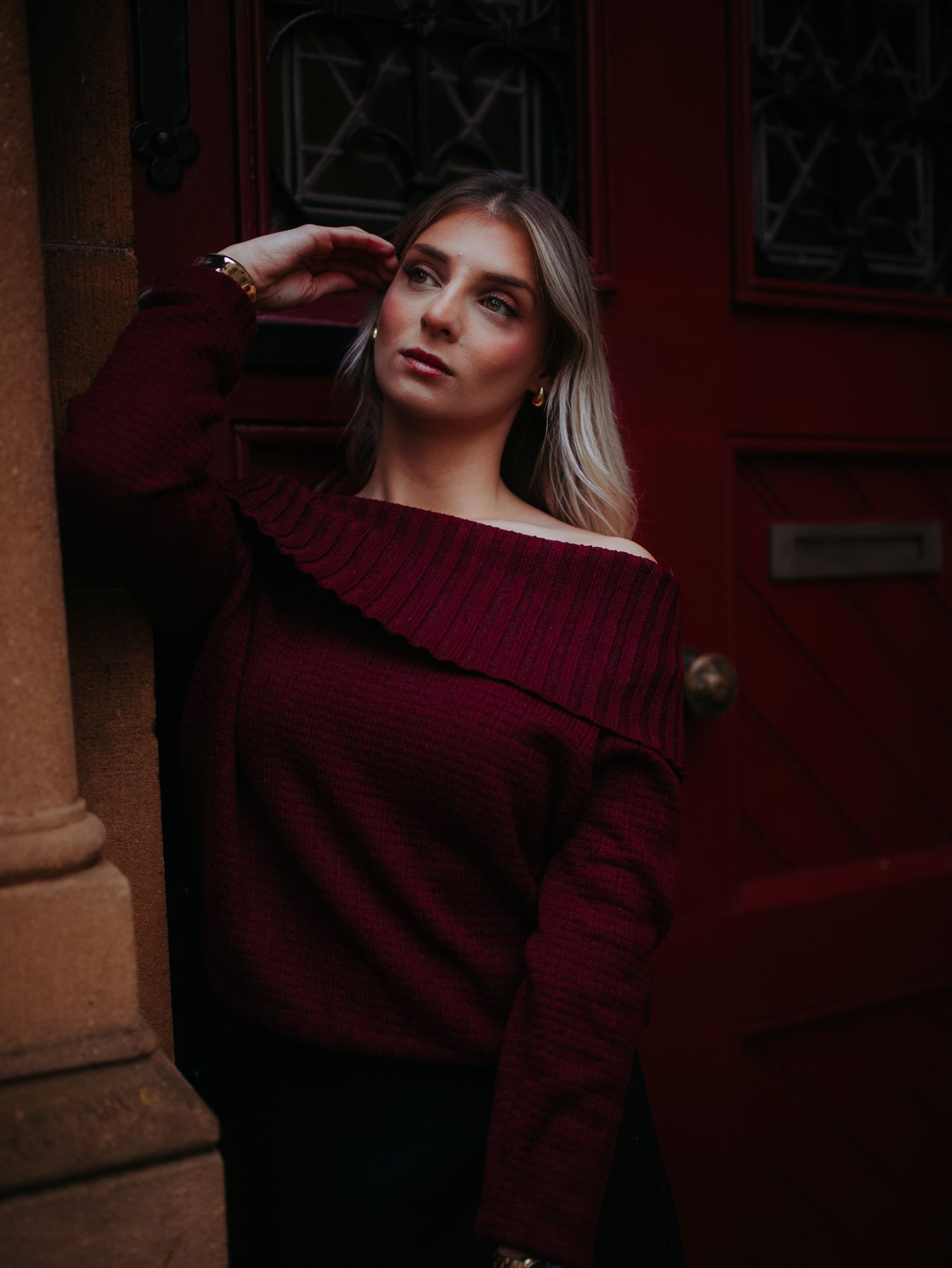 Blonde woman in off-shoulder burgundy knit sweater poses elegantly against a dark red vintage door.