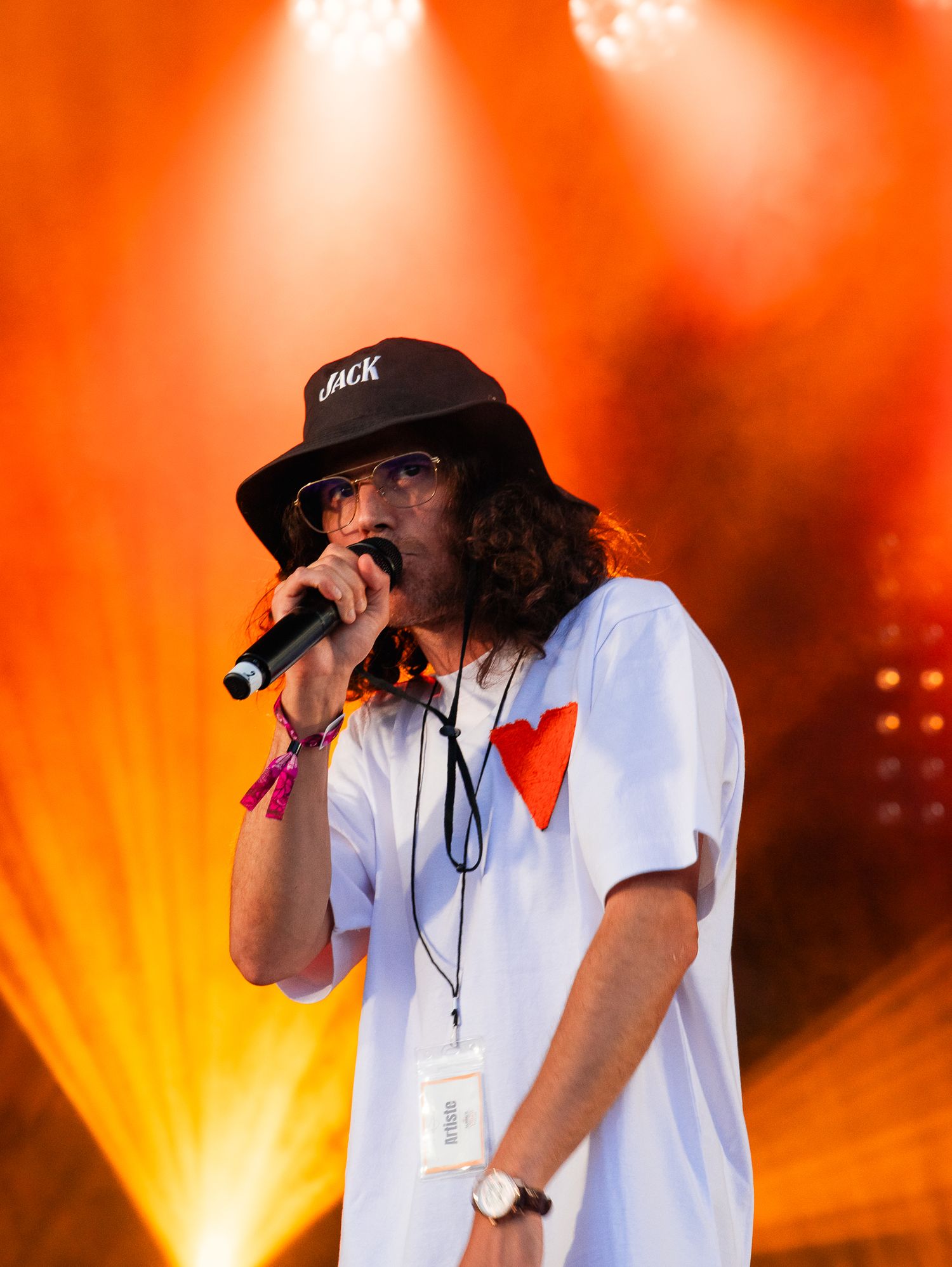 Un rappeur masculin se produisant sur scène portant un t-shirt blanc et un chapeau seau noir sous des lumières de scène orange.