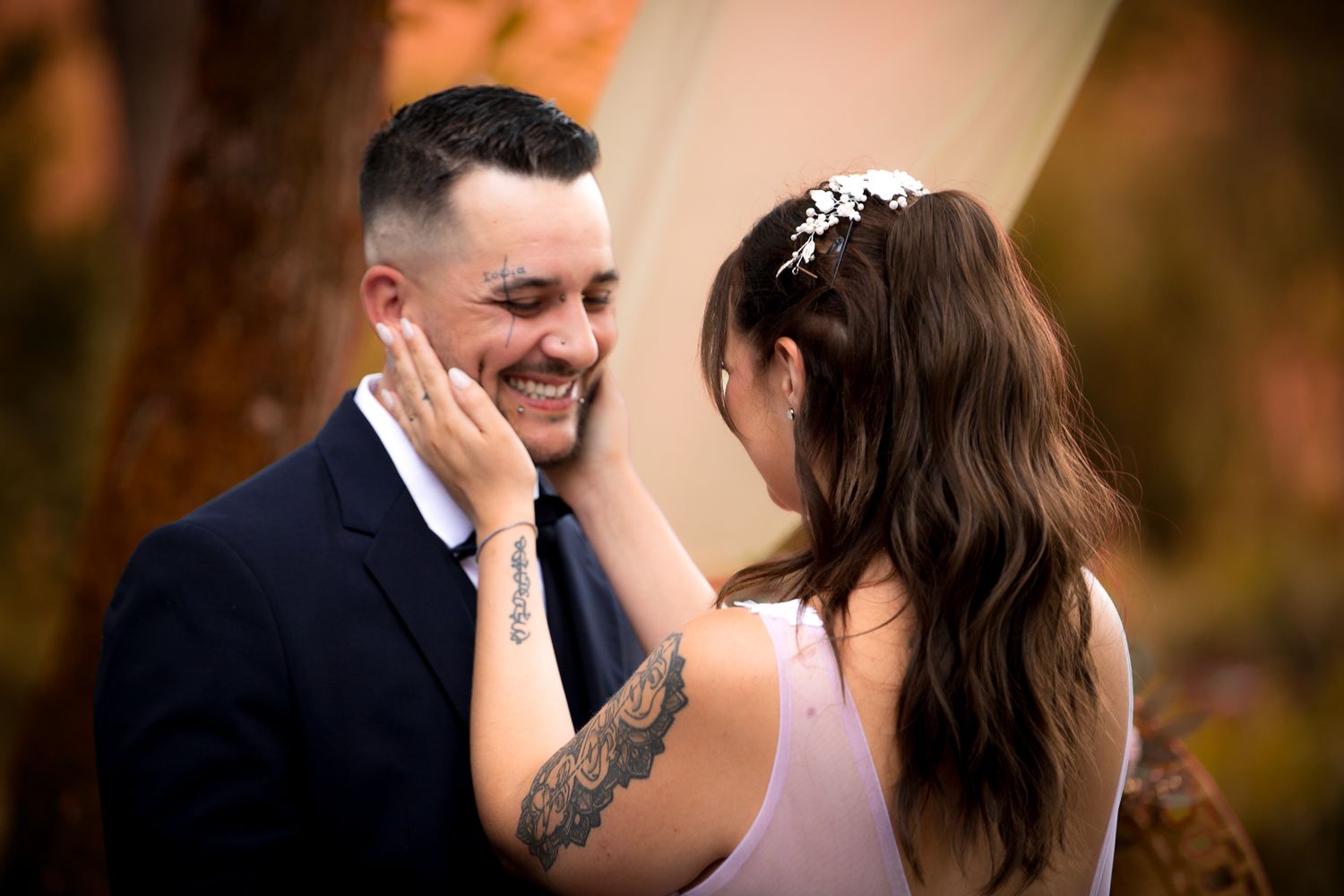 Groom smiling as tattooed bride tenderly touches his face during wedding ceremony.