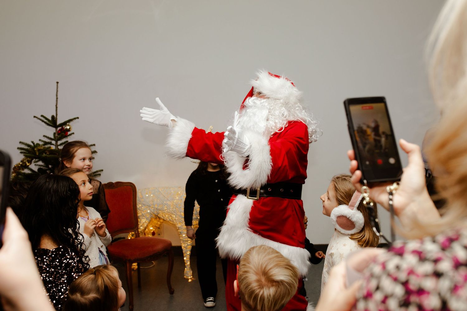 Santa Claus entertains children at a Christmas party while guests capture the moment on phones.