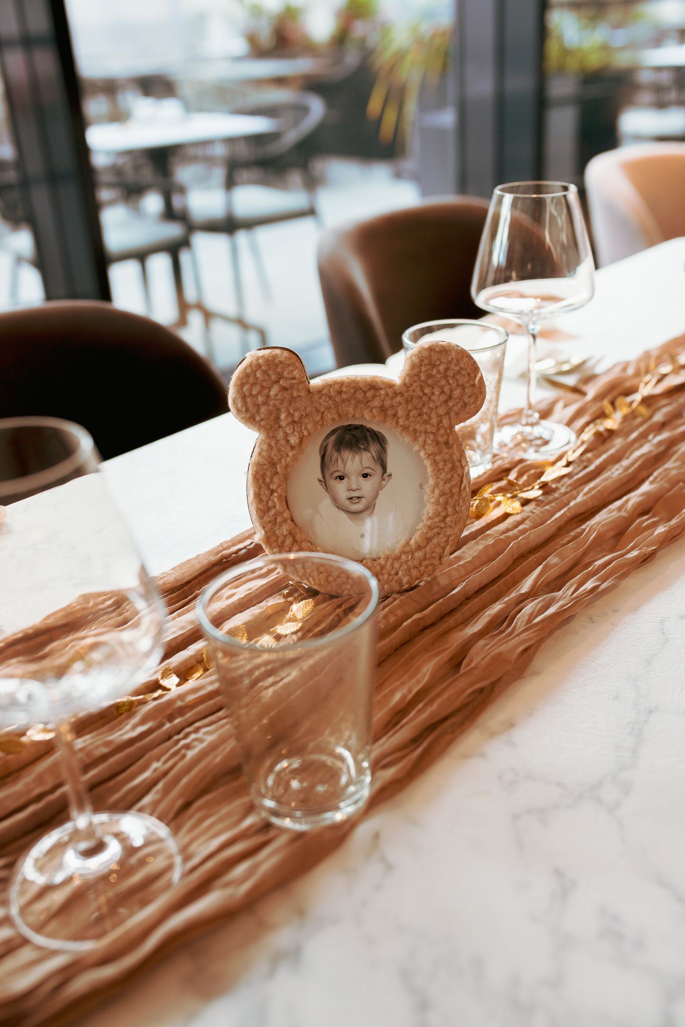 Child portrait in teddy bear frame displayed on marble table with wine glasses.