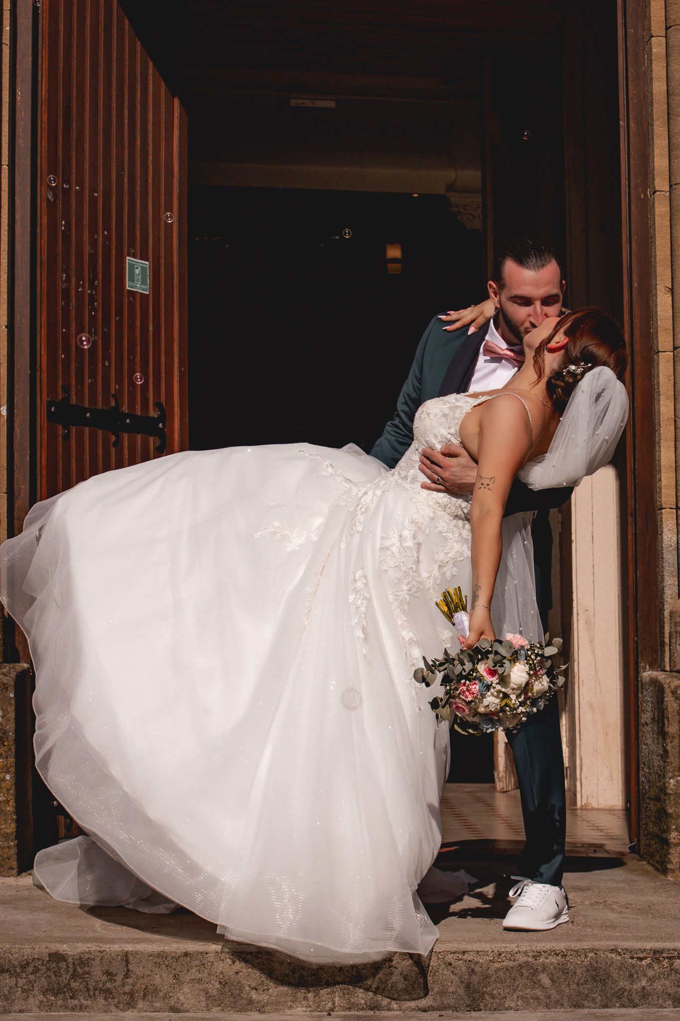 Bride and groom share a romantic kiss in a white lace wedding gown and teal suit at a church entrance.