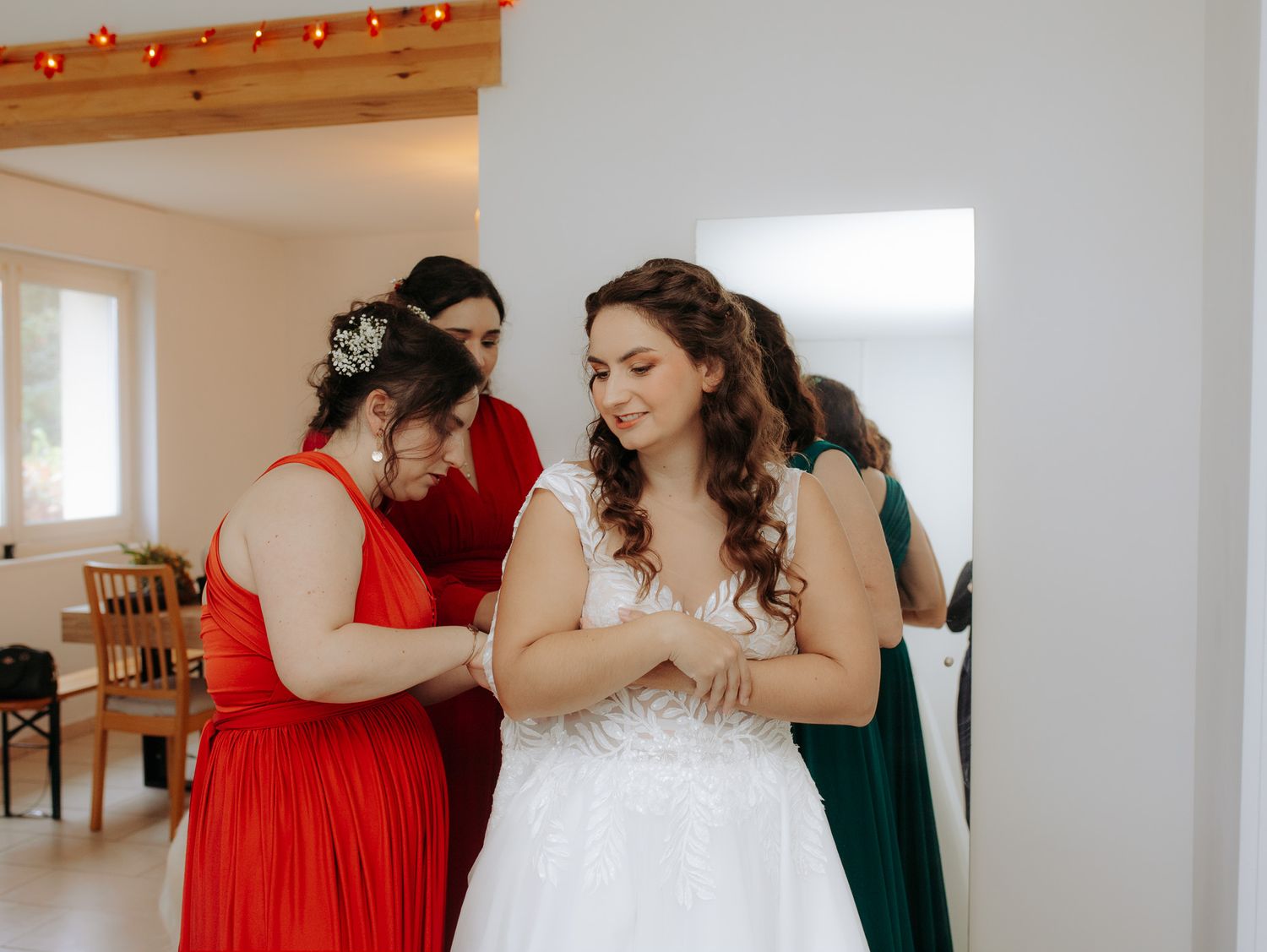 Bridesmaids in red and green dresses helping bride button her white lace wedding gown indoors.