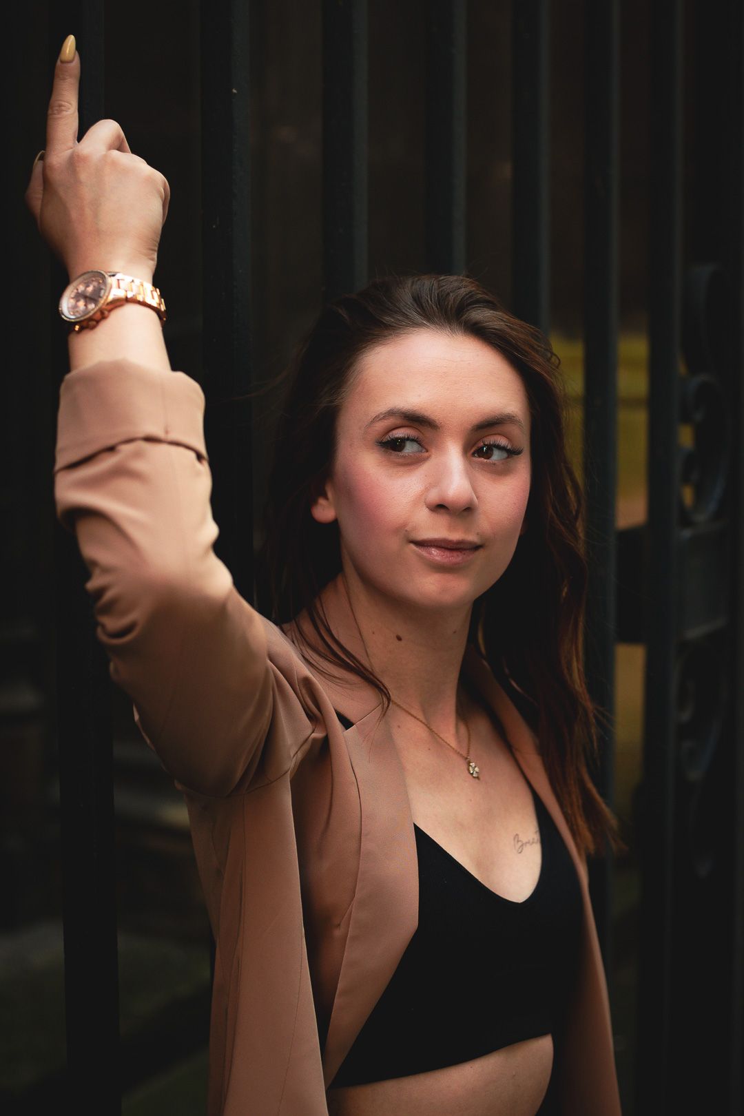 Brunette woman in beige blazer reaching up while posing against dark metal fence.