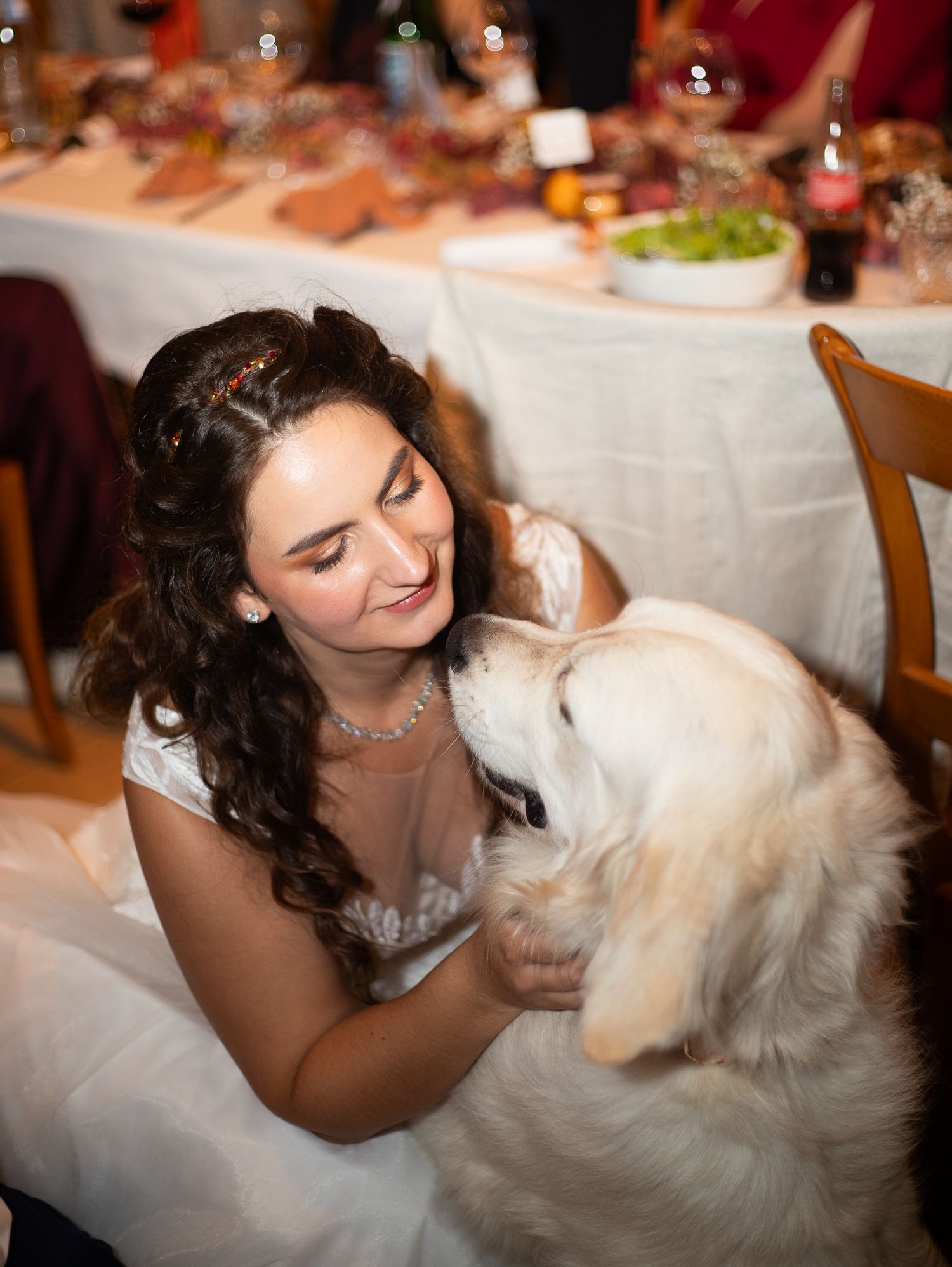 Bride in white dress smiling as a golden retriever dog licks her face at reception.