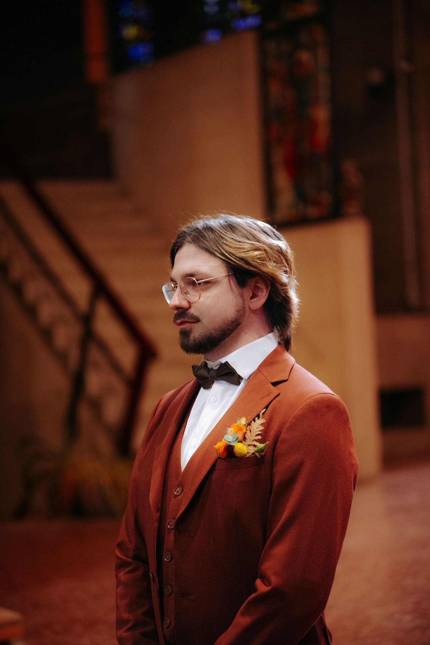 Elegant groom in a rust-colored velvet suit with bow tie and floral boutonniere standing inside a church.