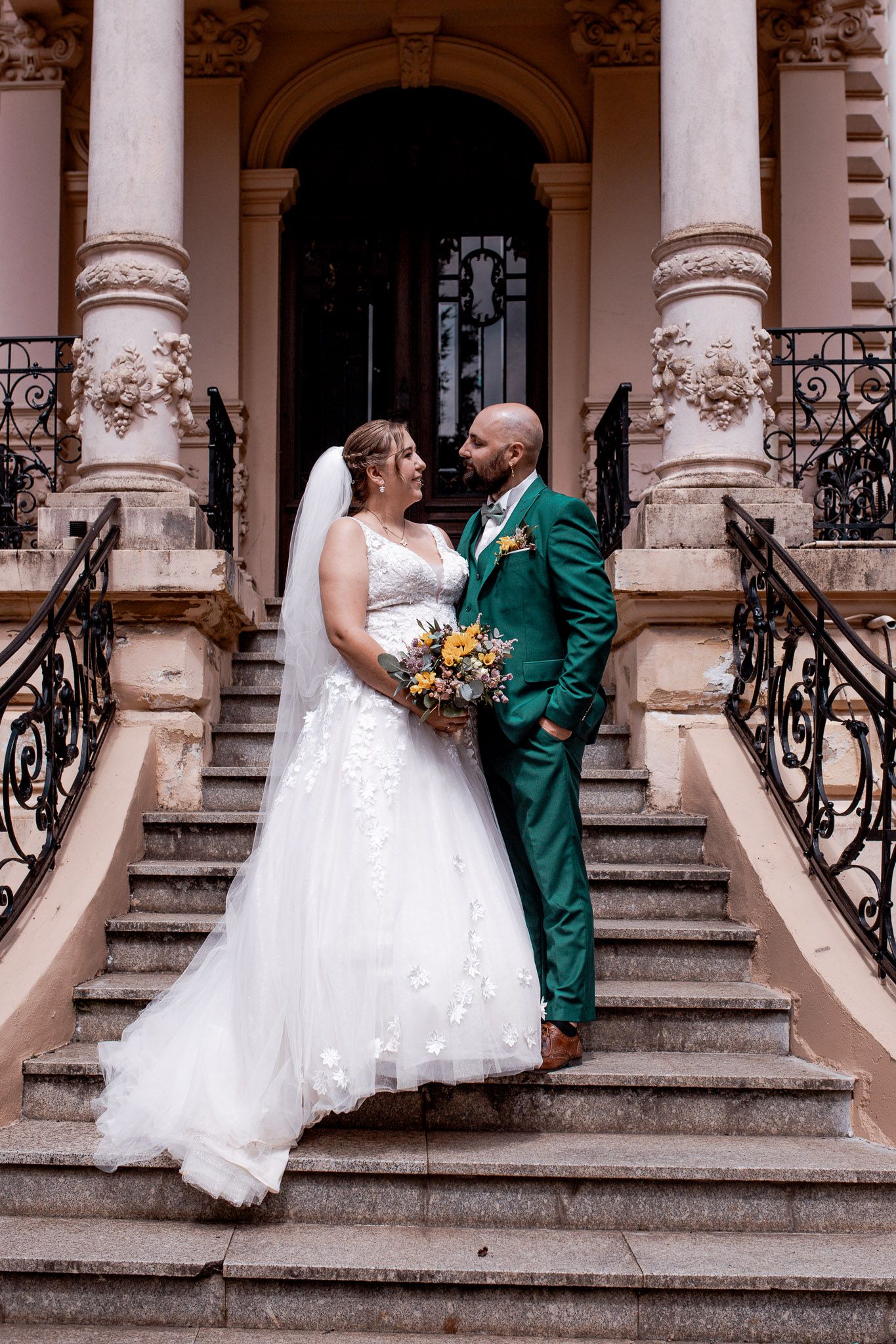 Bride and groom share a romantic moment on grand stone steps, she in white lace gown, he in emerald suit with bouquet.