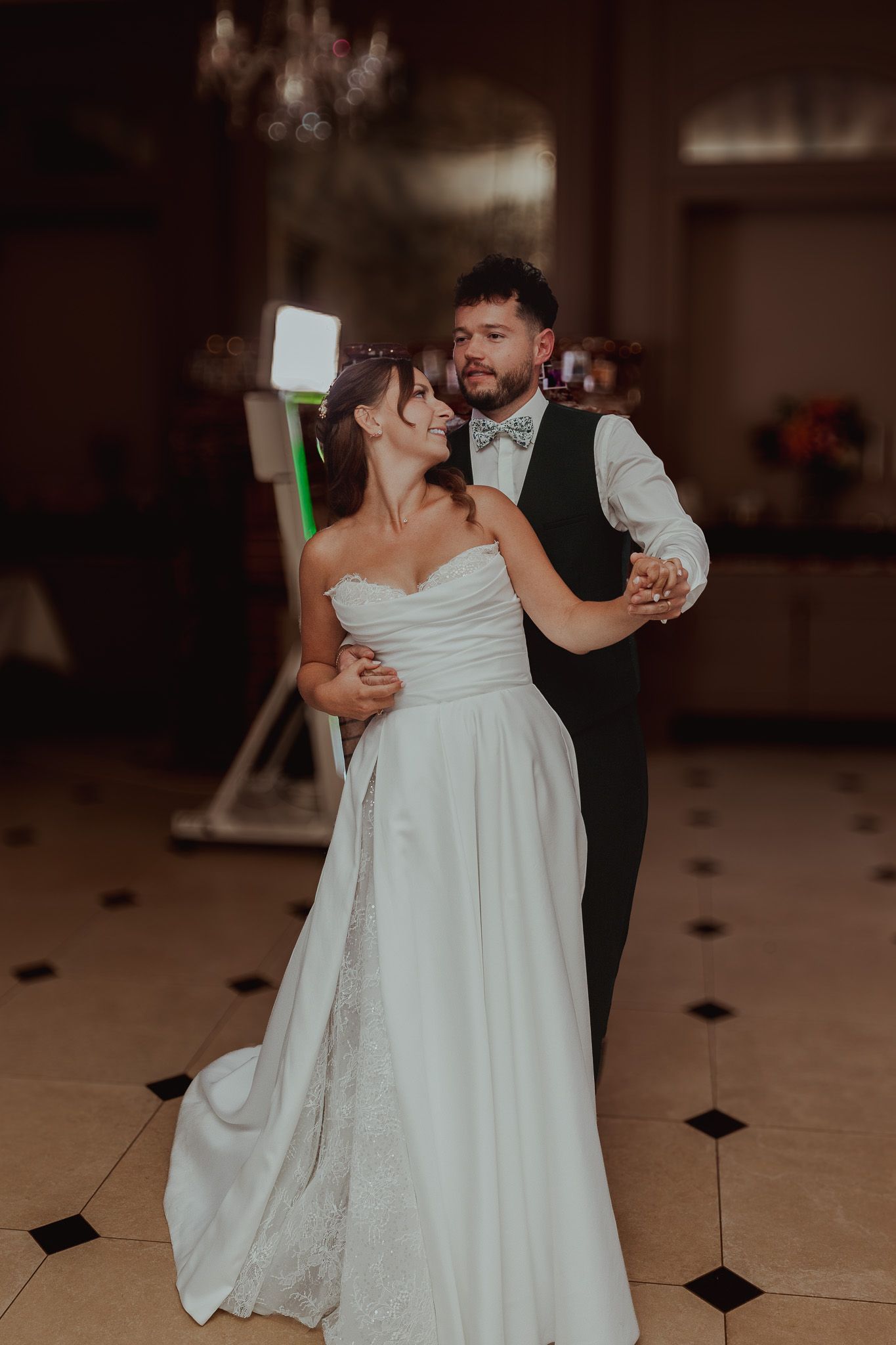 Bride and groom share their first dance, she in a strapless white gown, he in a vest and bow tie, at their wedding reception.