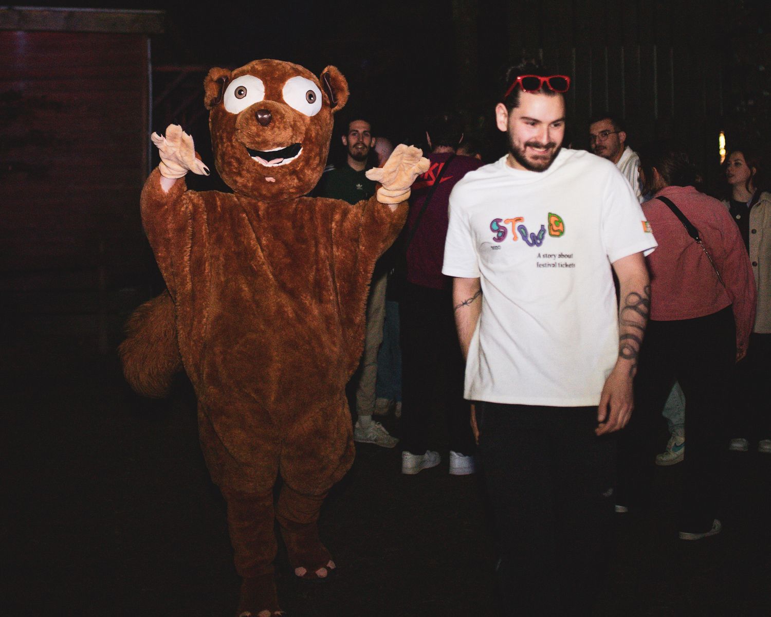 Man in STWS t-shirt posing and laughing beside a brown bear mascot costume at an indoor event.