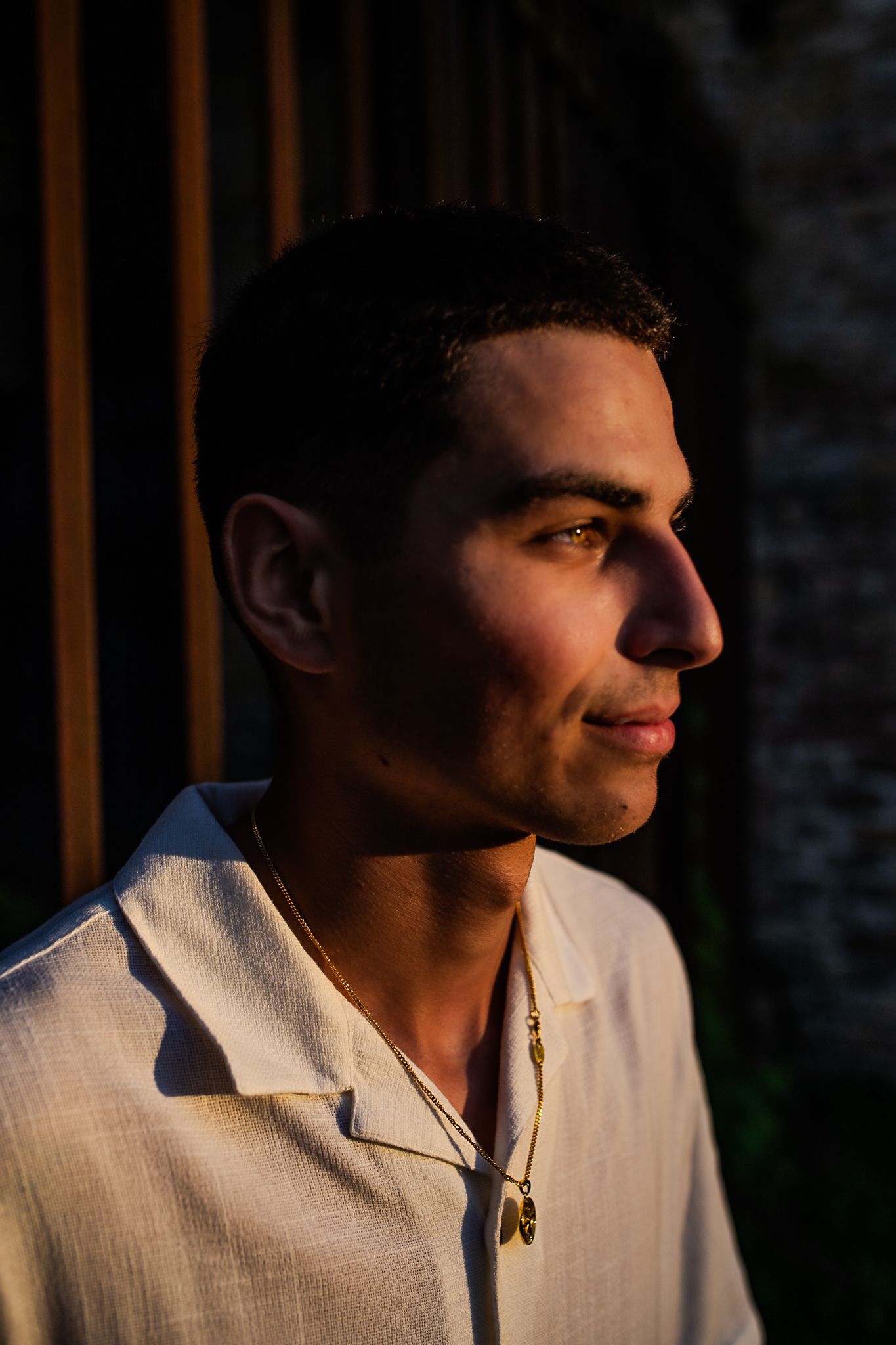 Young man in white linen shirt wearing gold necklace, bathed in warm golden hour sunlight, profile portrait.
