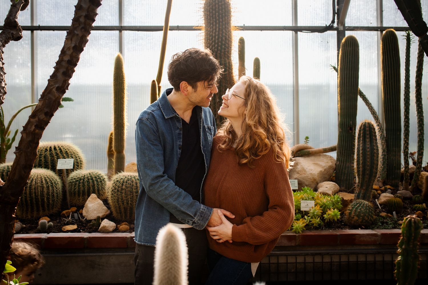 Romantic couple gazing at each other tenderly surrounded by tall cacti in a bright greenhouse setting.