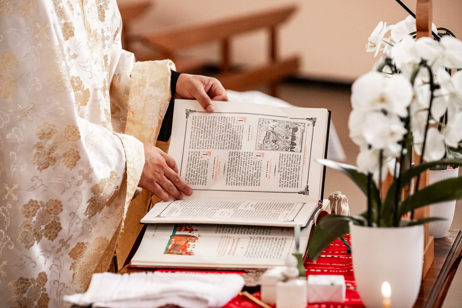 Priest in golden vestments reading from a liturgical book beside white orchids during a baptism.