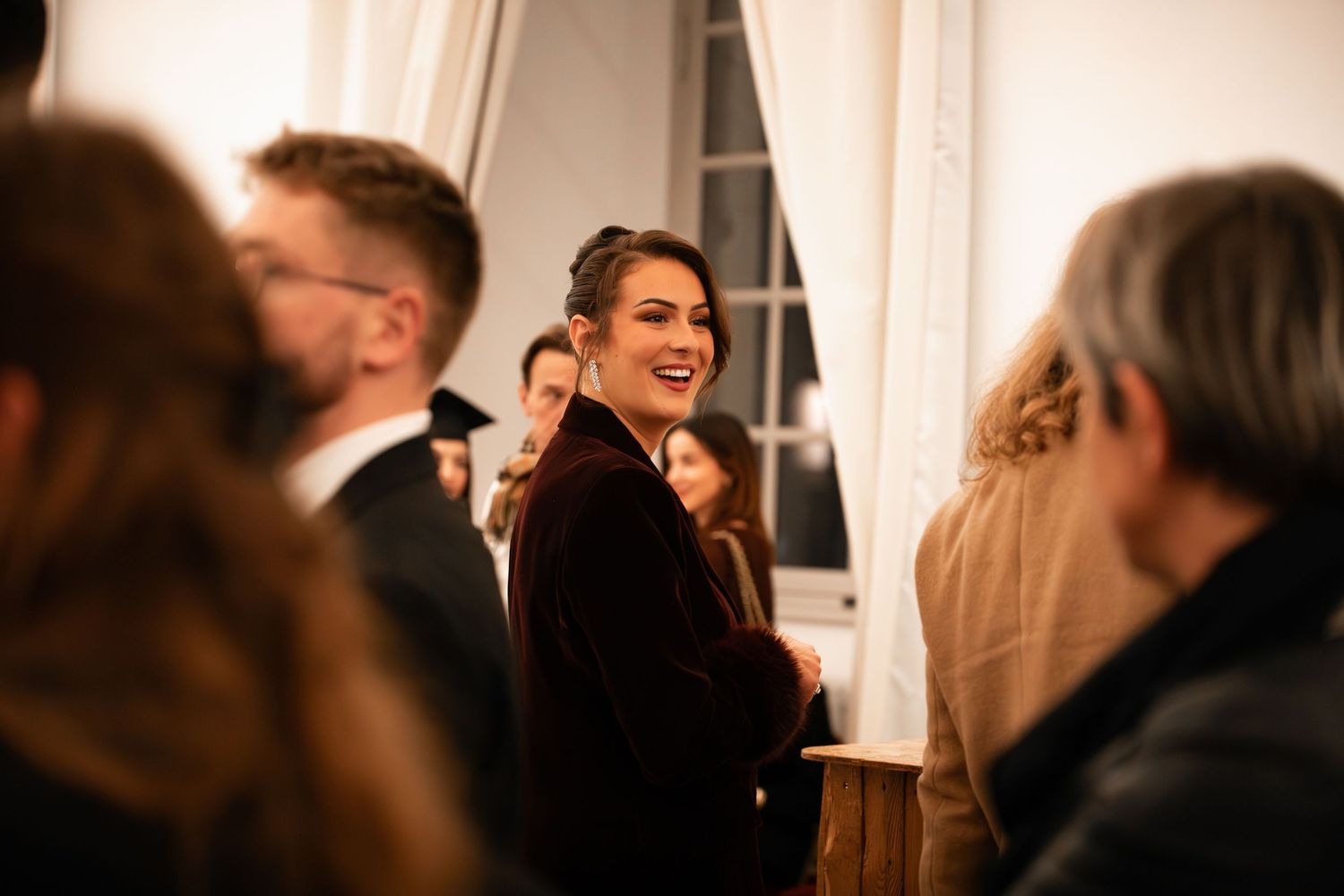 Smiling woman in burgundy velvet blazer socializing at indoor evening event.