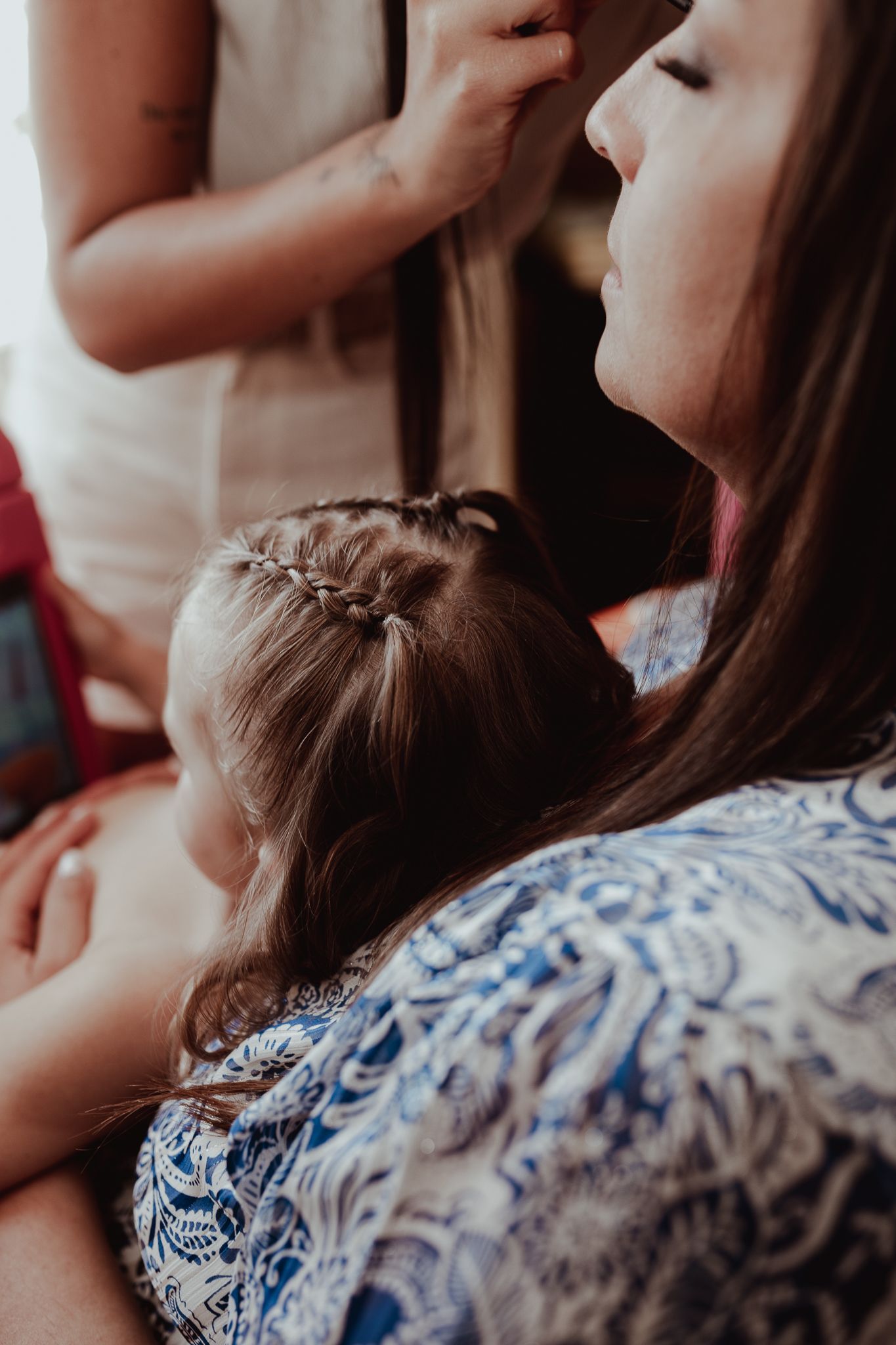 Young flower girl with braided hair sits on bride's lap during wedding hair preparation.