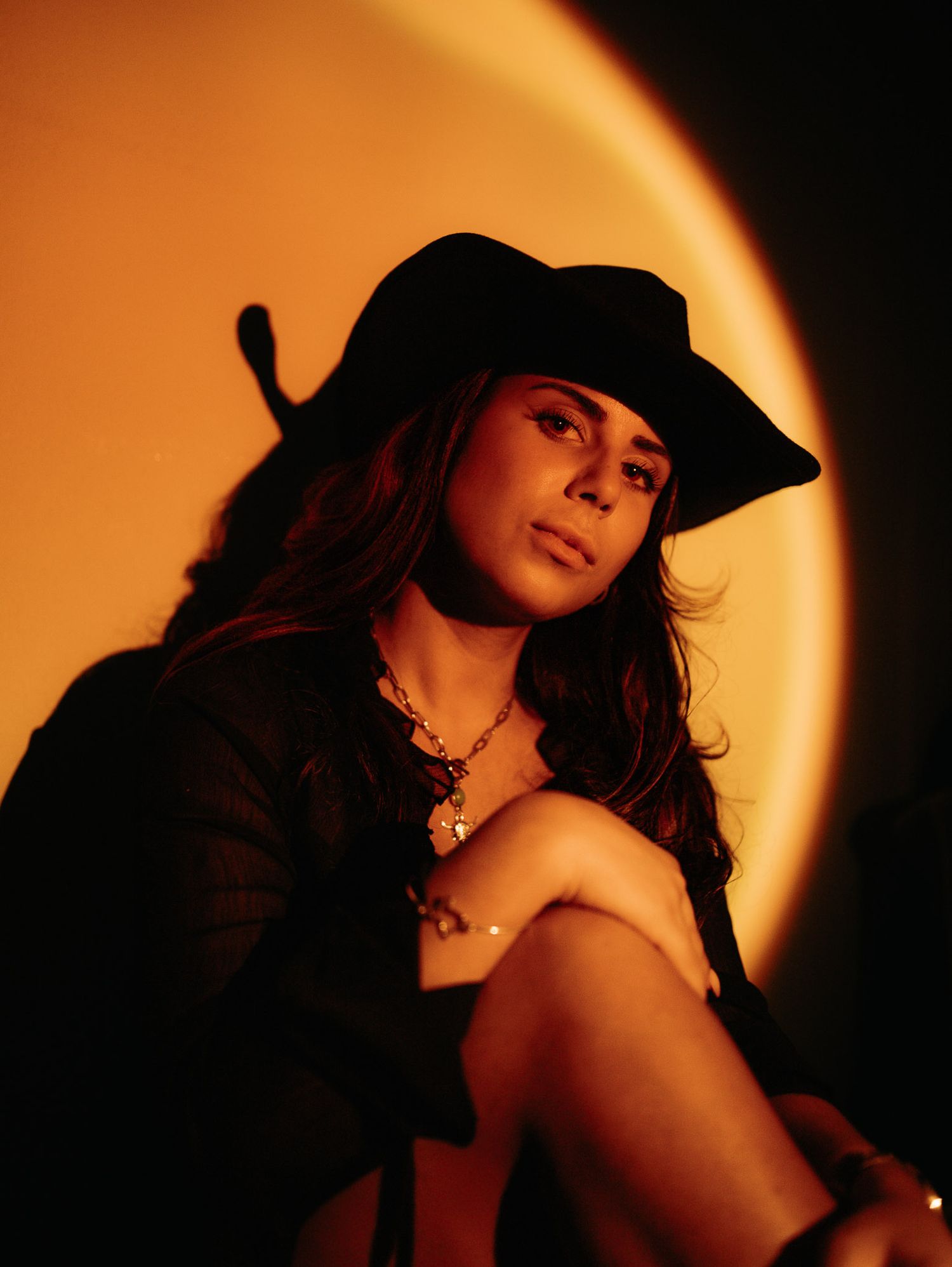 Woman in black hat sitting against warm orange sunset lamp glow background.