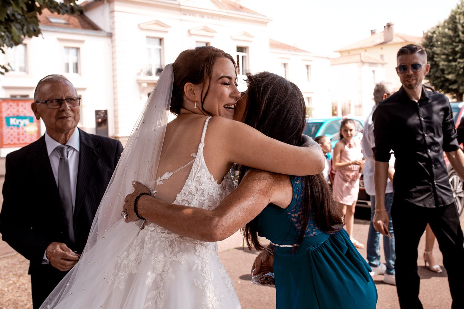 Bride in white gown receiving a warm hug from a guest in a teal dress at an outdoor wedding.