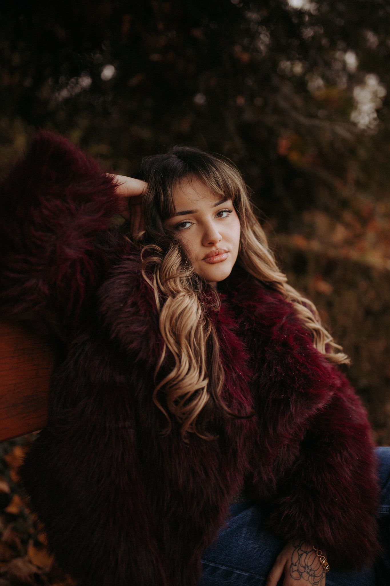 Young woman in burgundy faux fur coat with wavy blonde hair poses outdoors in autumn setting.