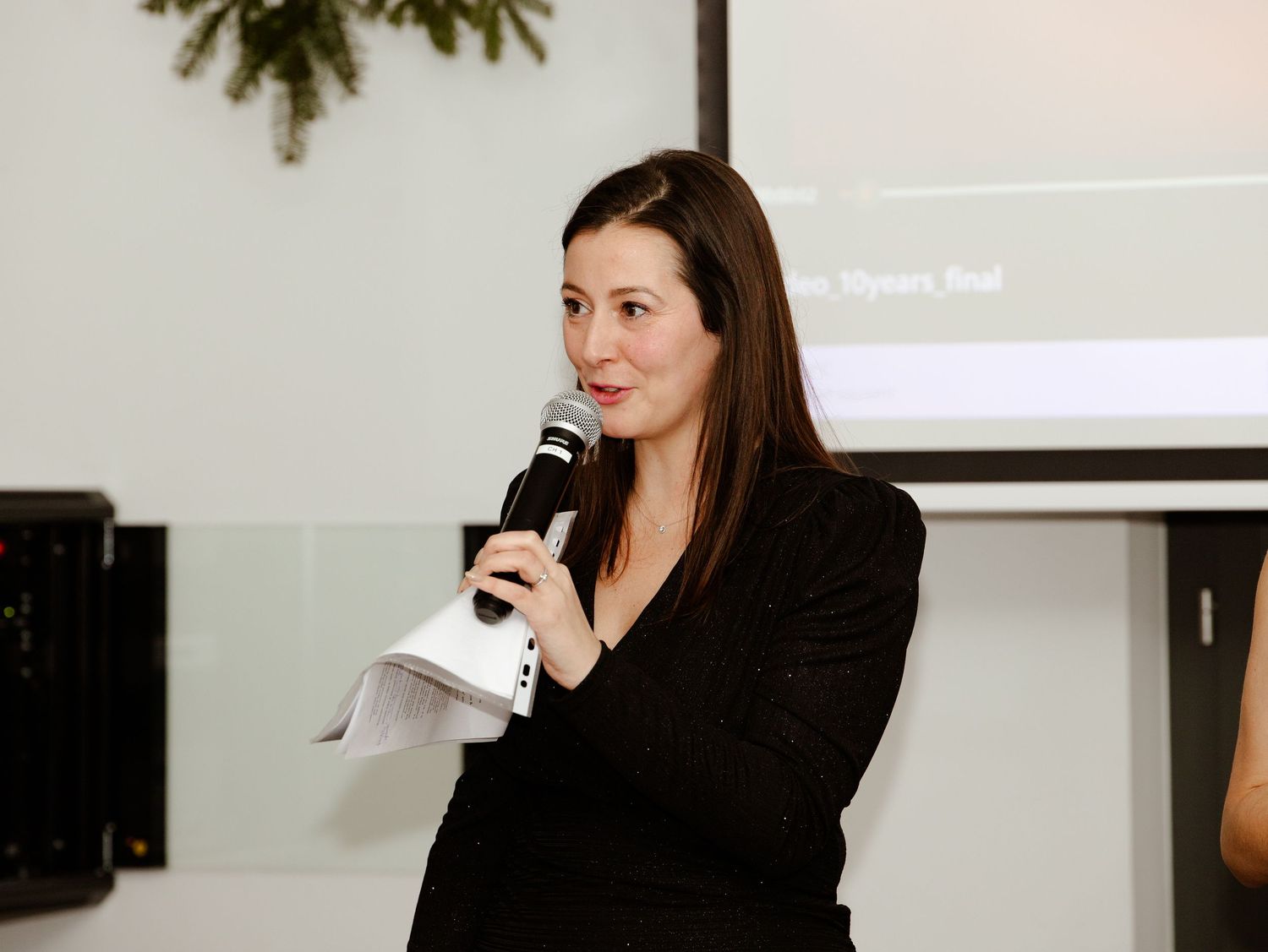 A woman in a black blazer speaks into a microphone during a professional presentation or event.