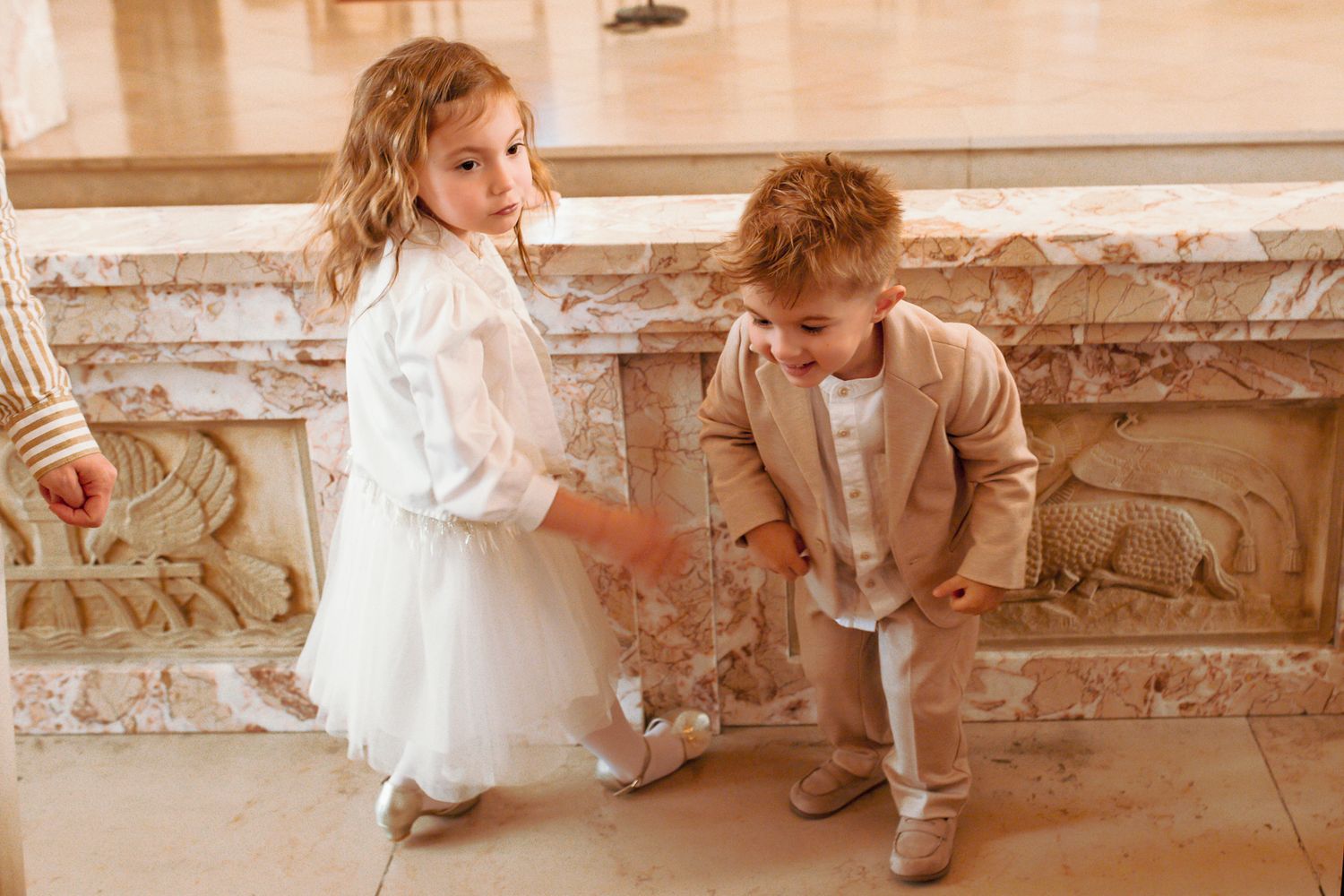 Two young children dressed in formal attire standing near ornate stone decor at a baptism.